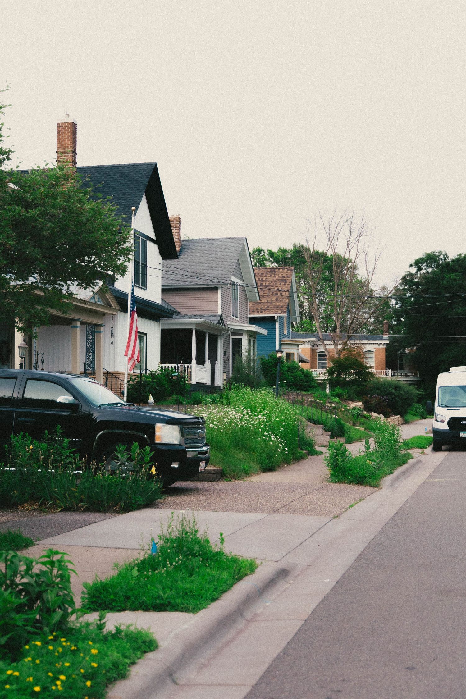 A quiet residential street lined with historic homes, lush greenery, parked trucks, and an American flag on a cloudy day.