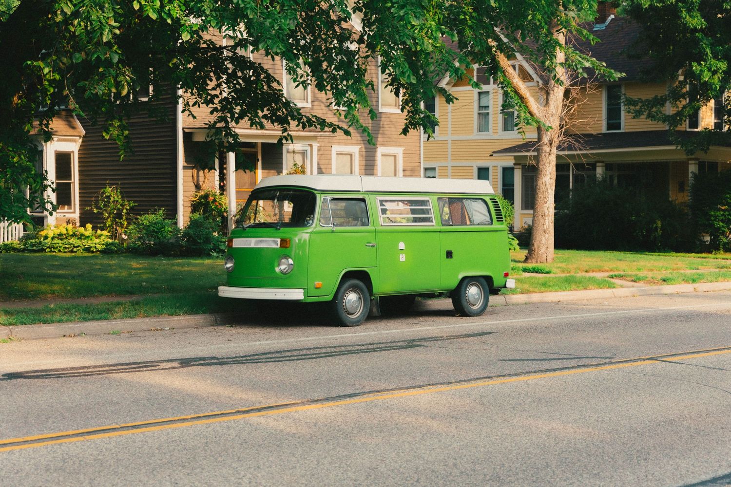 Vintage green Volkswagen Type 2 bus parked on a quiet suburban street lined with trees and classic homes.