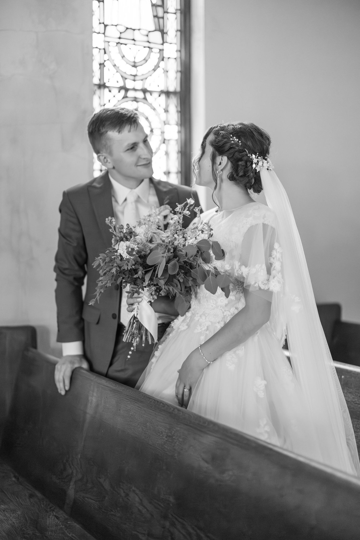 Black and white photo of bride and groom sharing a tender moment in a church pew with stained glass window.