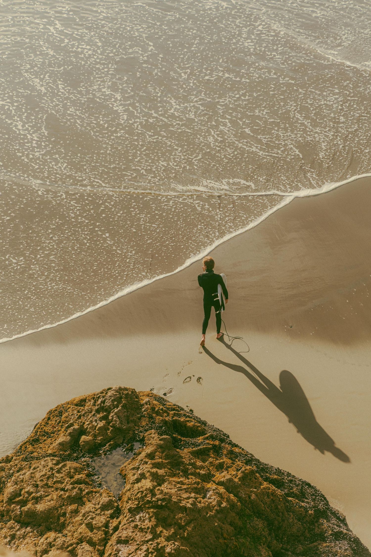 Aerial view of surfer in wetsuit carrying surfboard walking along sandy beach shoreline with ocean waves.