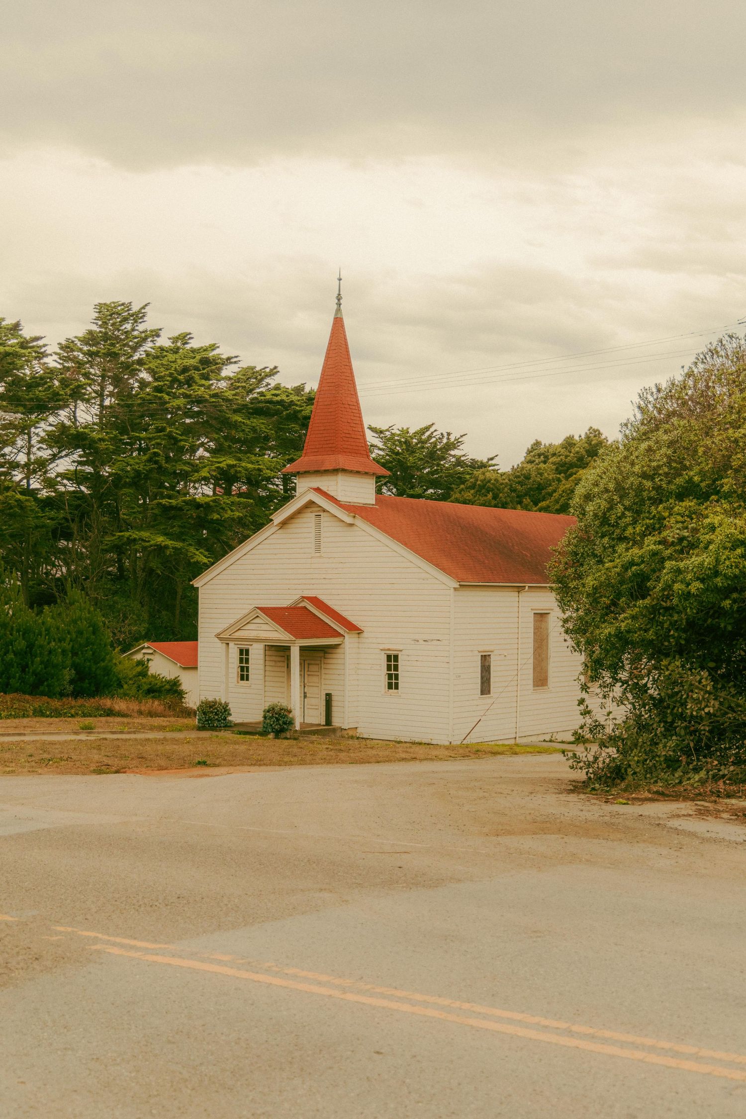 Charming white wooden church with a red steeple and roof surrounded by lush green trees under an overcast sky.