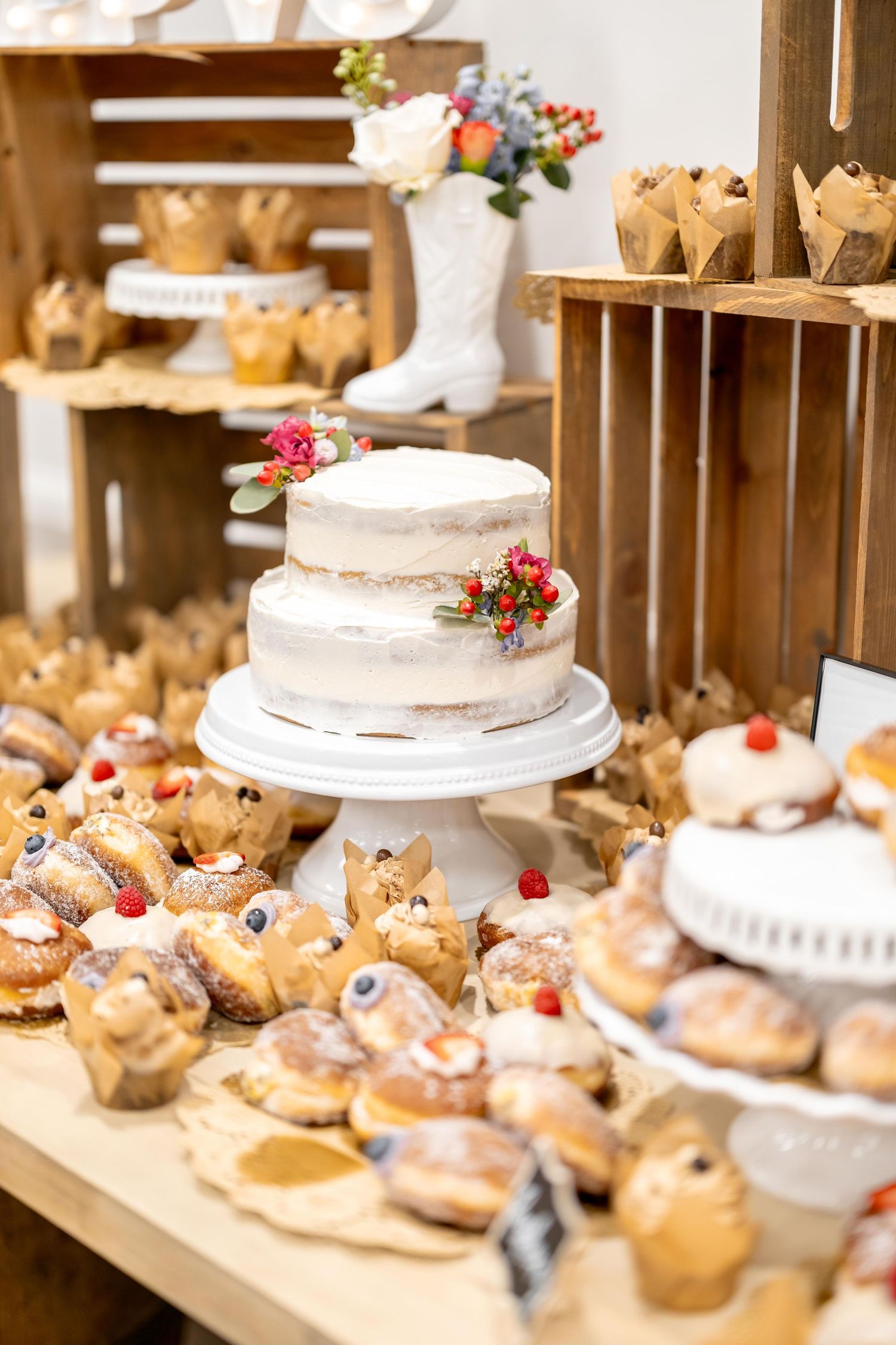 Rustic wedding dessert table featuring naked cake with floral decor surrounded by pastries and cupcakes on wooden shelves.