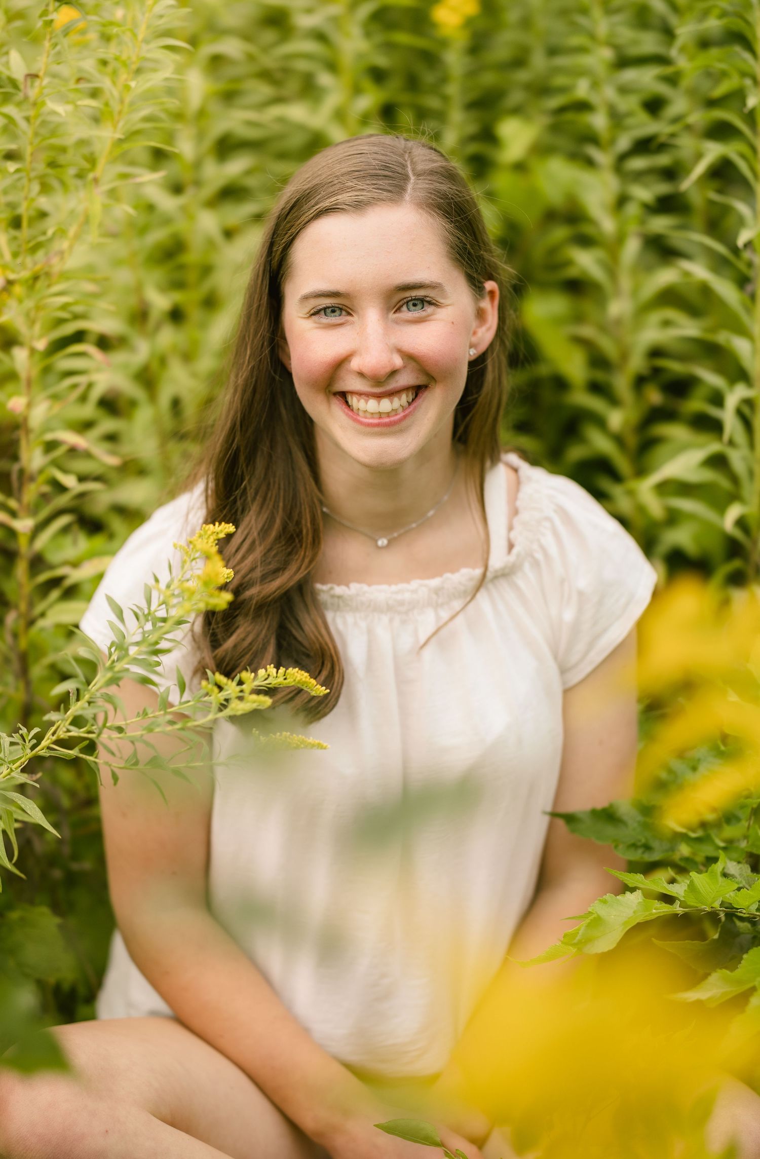 Happy teenage girl in white top sitting among yellow wildflowers smiling for senior portrait photography.