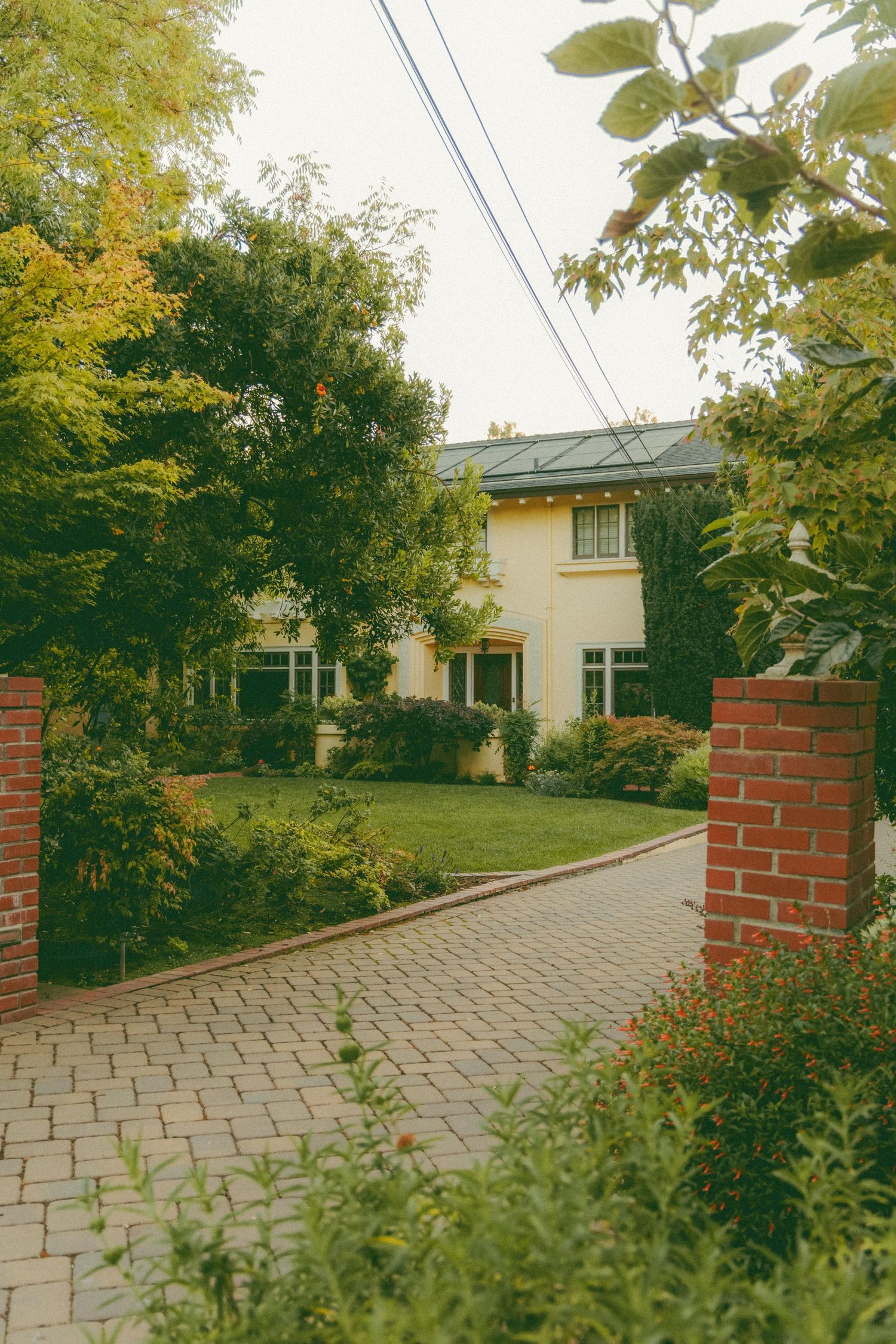 Charming yellow house with brick pillars, cobblestone driveway, and lush garden landscaping.