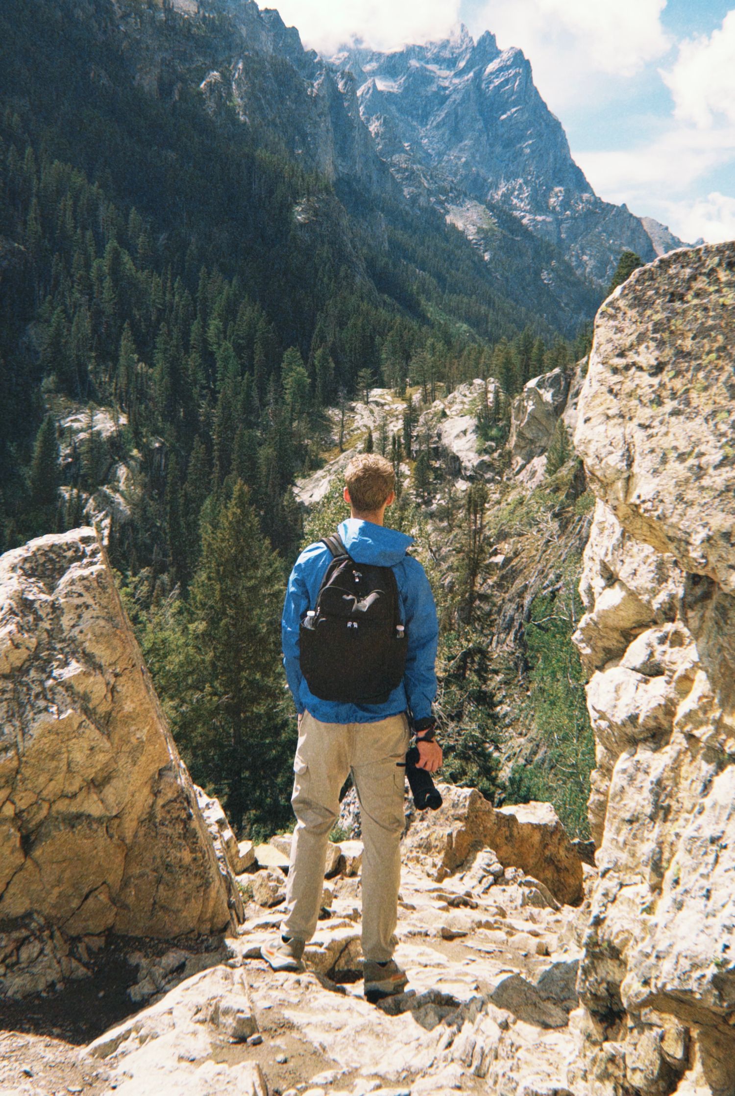 Hiker with black backpack and camera stands on rocky overlook surveying forested mountain valley.