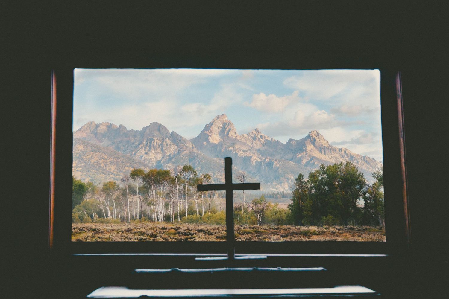 A wooden cross silhouetted against a scenic mountain landscape viewed through a rustic window frame.