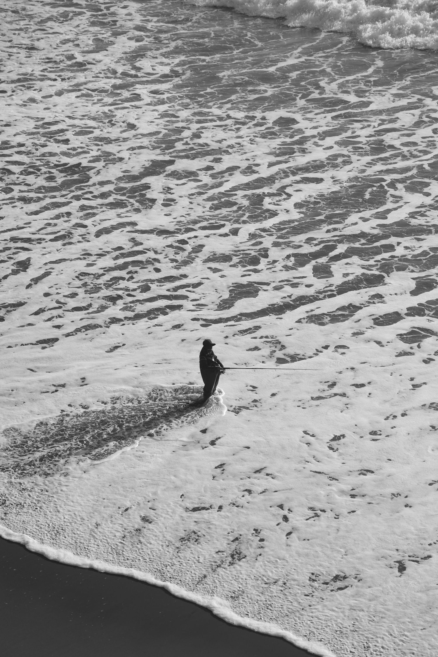 Aerial black and white photo of lone person standing at ocean shoreline with foamy waves on sandy beach.