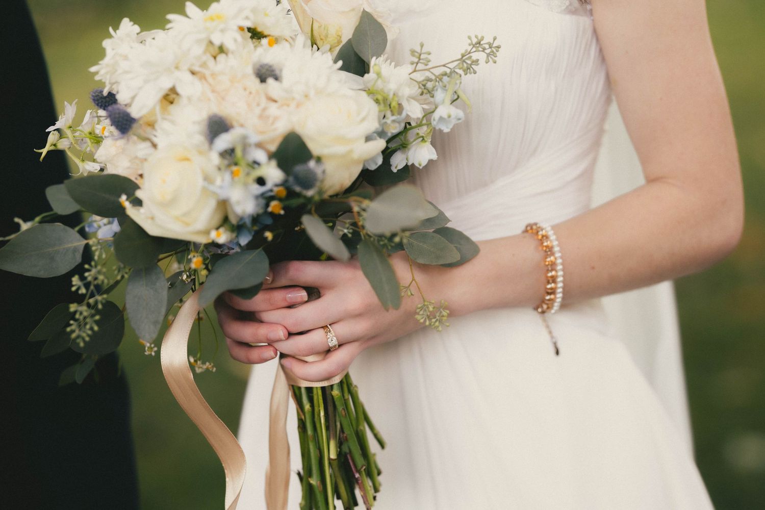 Bride holding white floral wedding bouquet with eucalyptus, wearing gold bracelet and wedding ring.