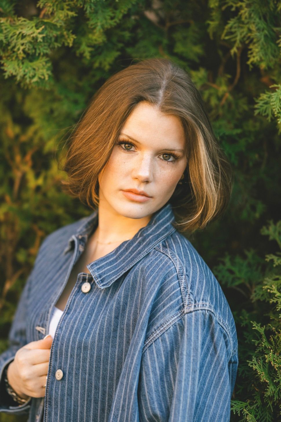 Young woman with bob haircut in striped denim jacket posing outdoors against lush green evergreen shrubs.