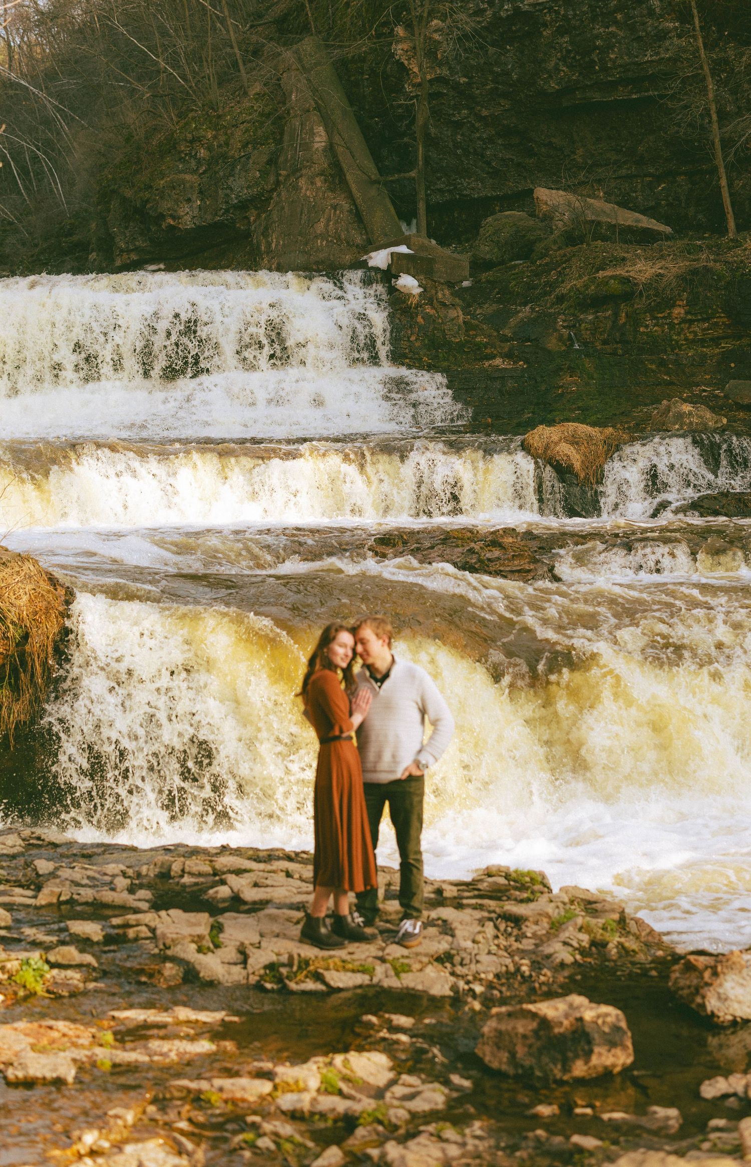 Couple embracing romantically in front of a cascading waterfall on a rocky riverbank.