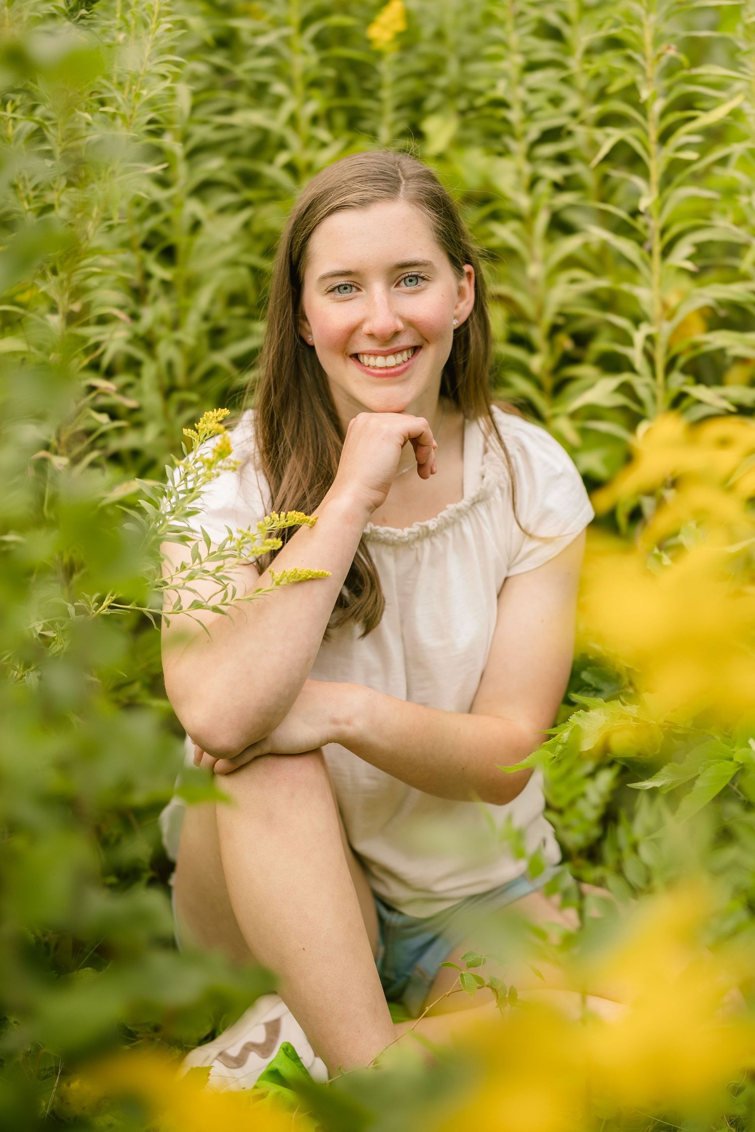 Smiling young woman sitting among lush green plants and yellow wildflowers, wearing a white top and denim shorts.