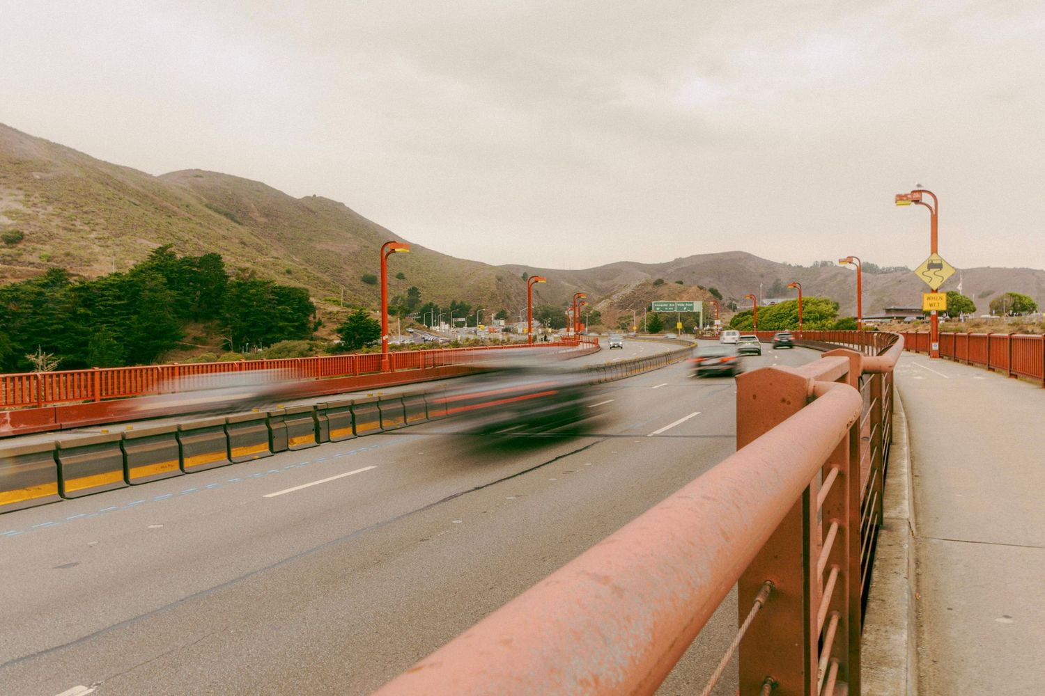 Long exposure shot of cars crossing a red bridge with mountains and overcast sky in urban landscape.