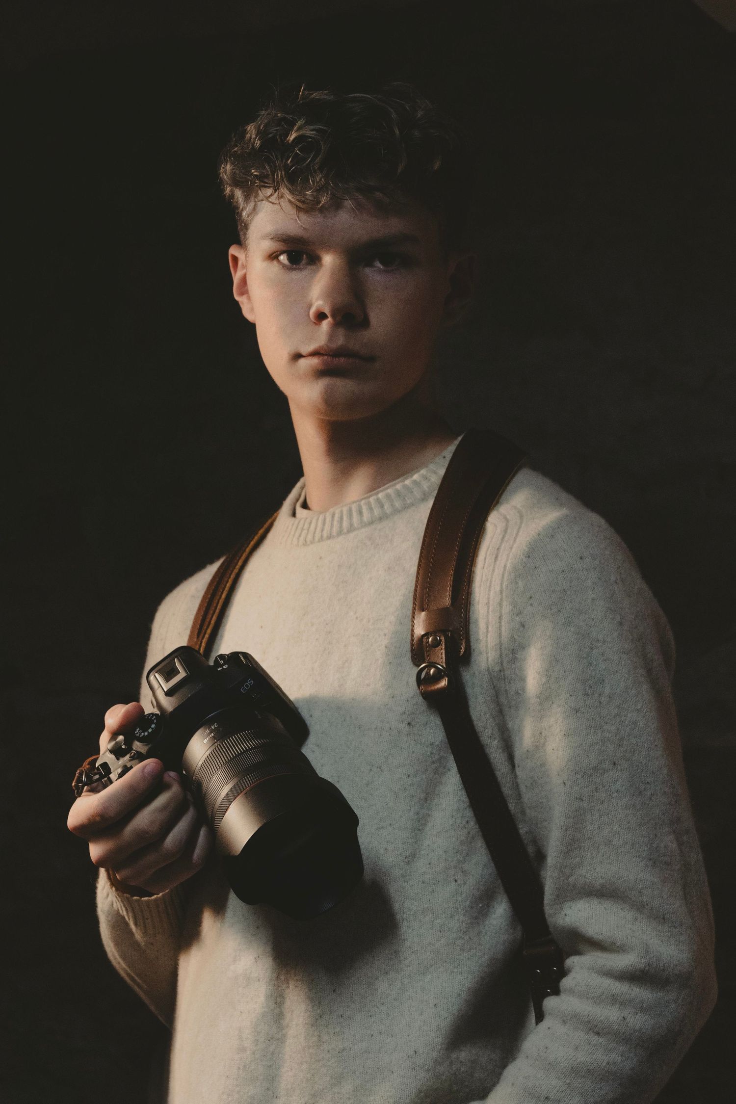 Young male photographer in white sweater holding DSLR camera with leather strap in moody dark lighting in Stillwater Minnesota