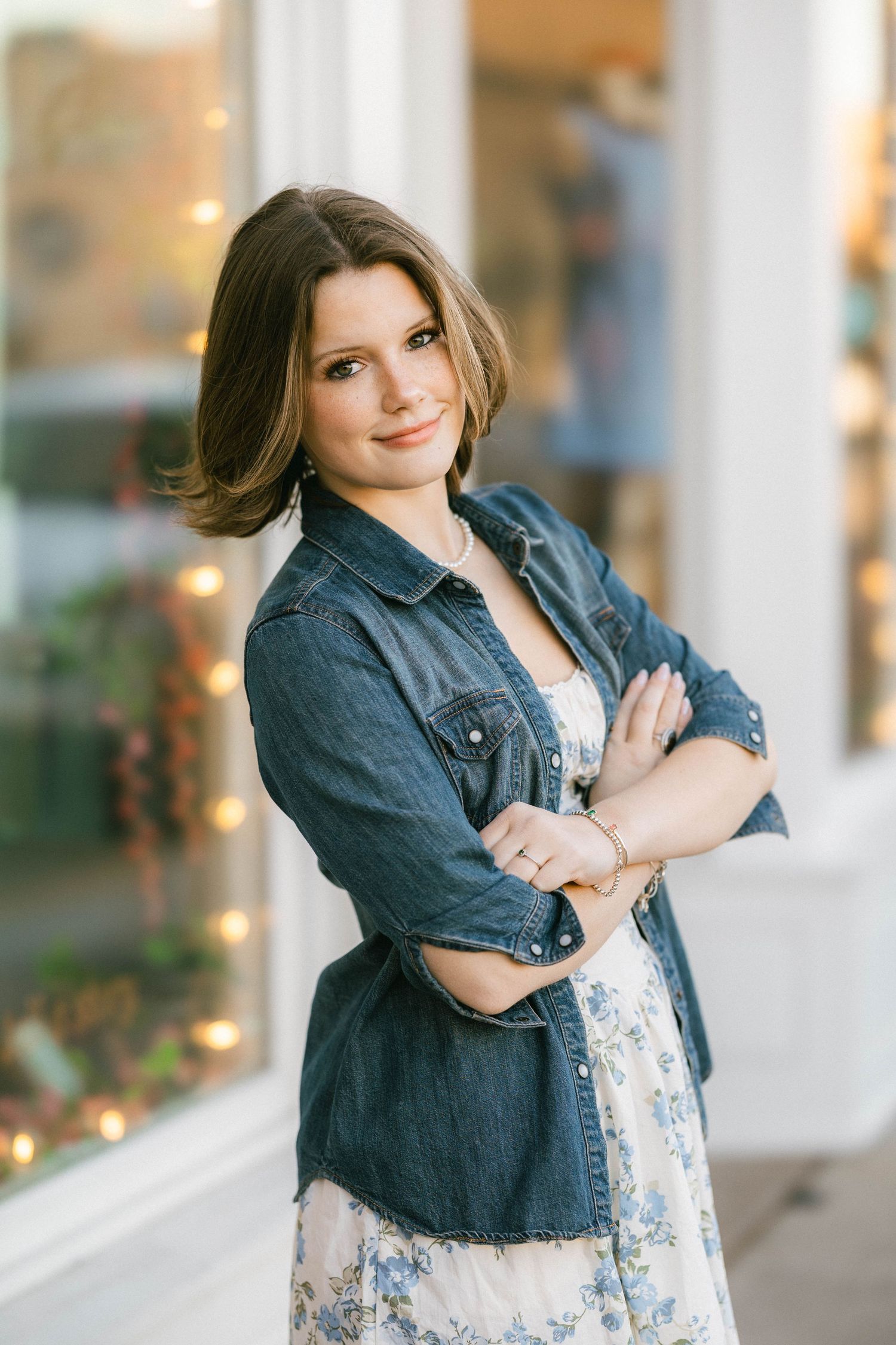 A young woman in a denim jacket and floral dress stands with arms crossed, smiling outdoors near a storefront.