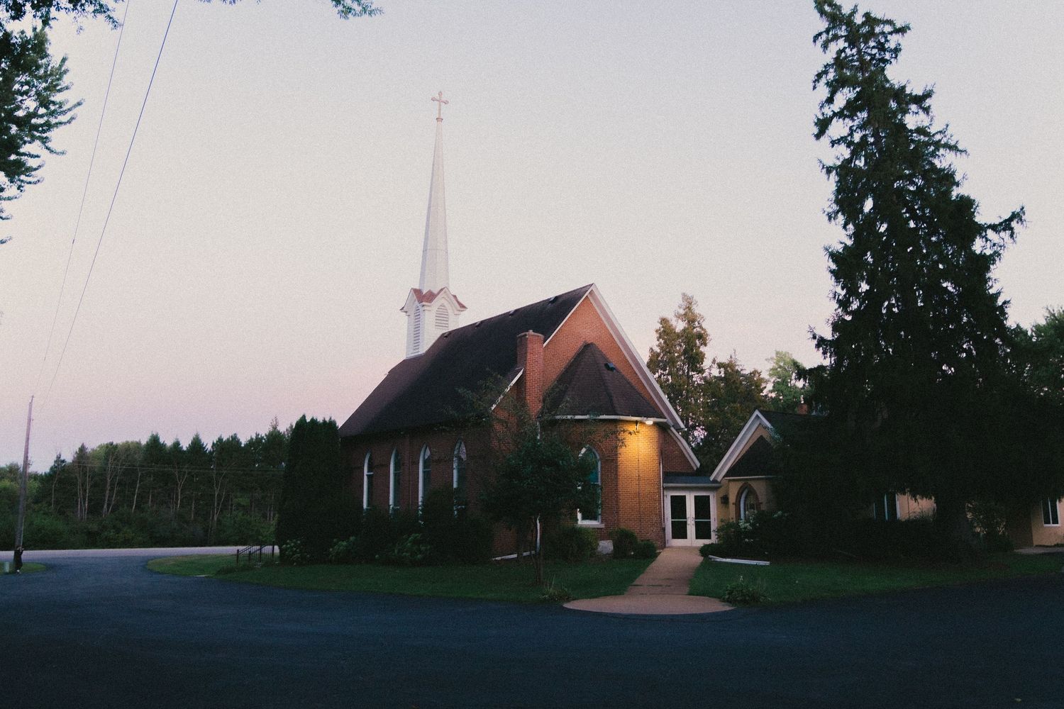 Small brick church with white steeple illuminated at dusk, surrounded by tall trees and a quiet rural road.