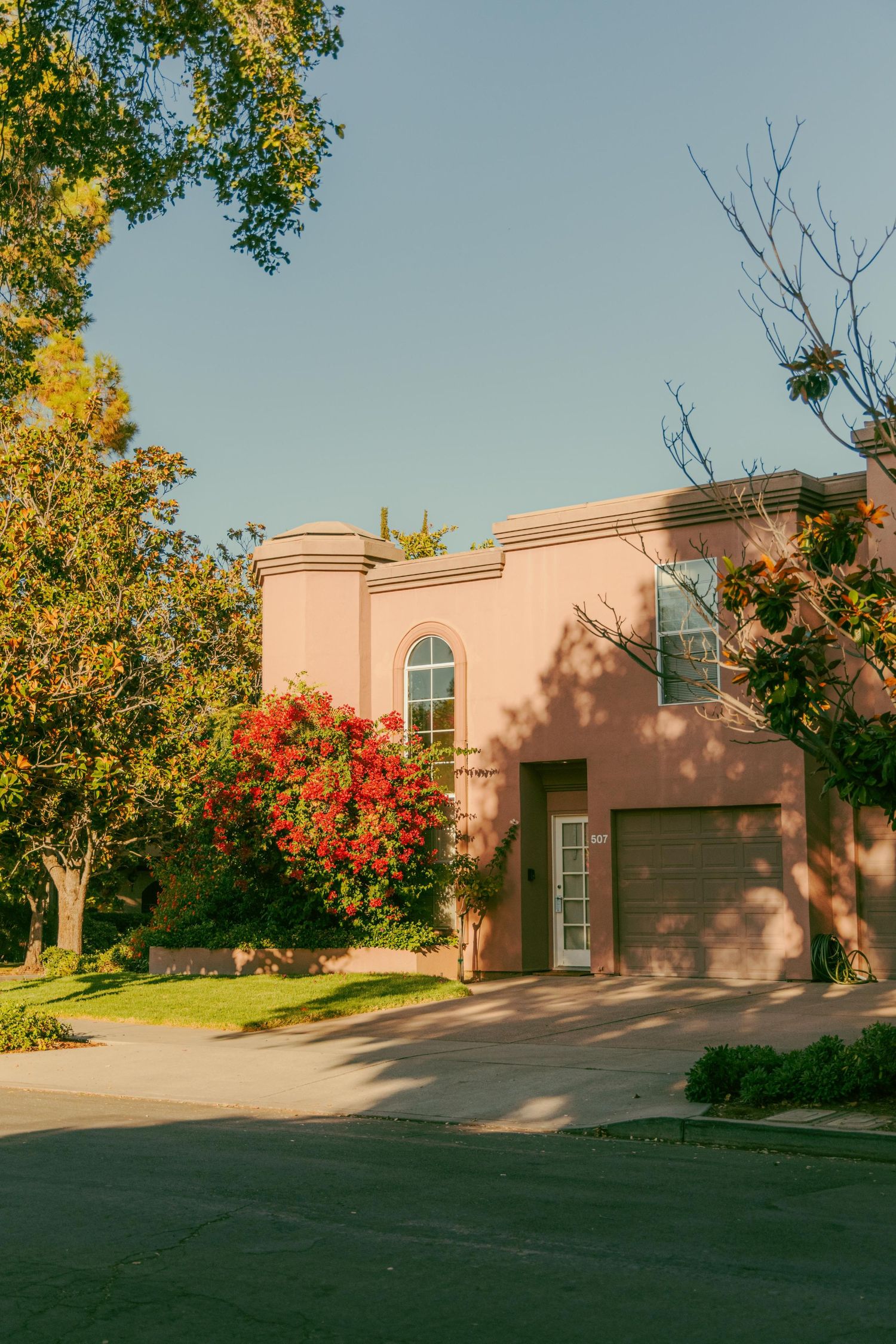 Pink stucco two-story home with arched windows and garage surrounded by colorful trees in warm sunlight.