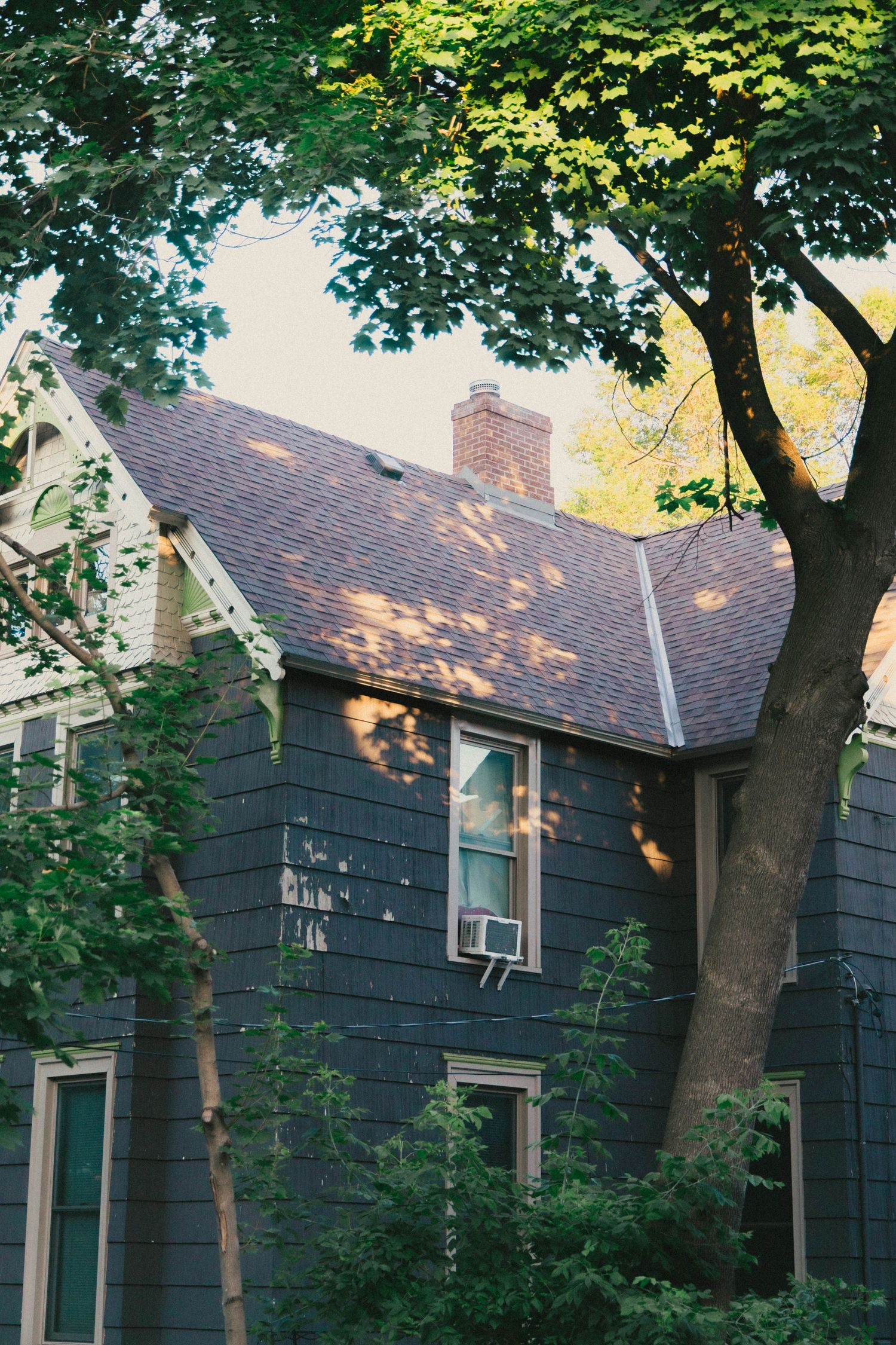 Dark gray clapboard house with brick chimney, shingle roof, and window AC unit, framed by lush green trees.