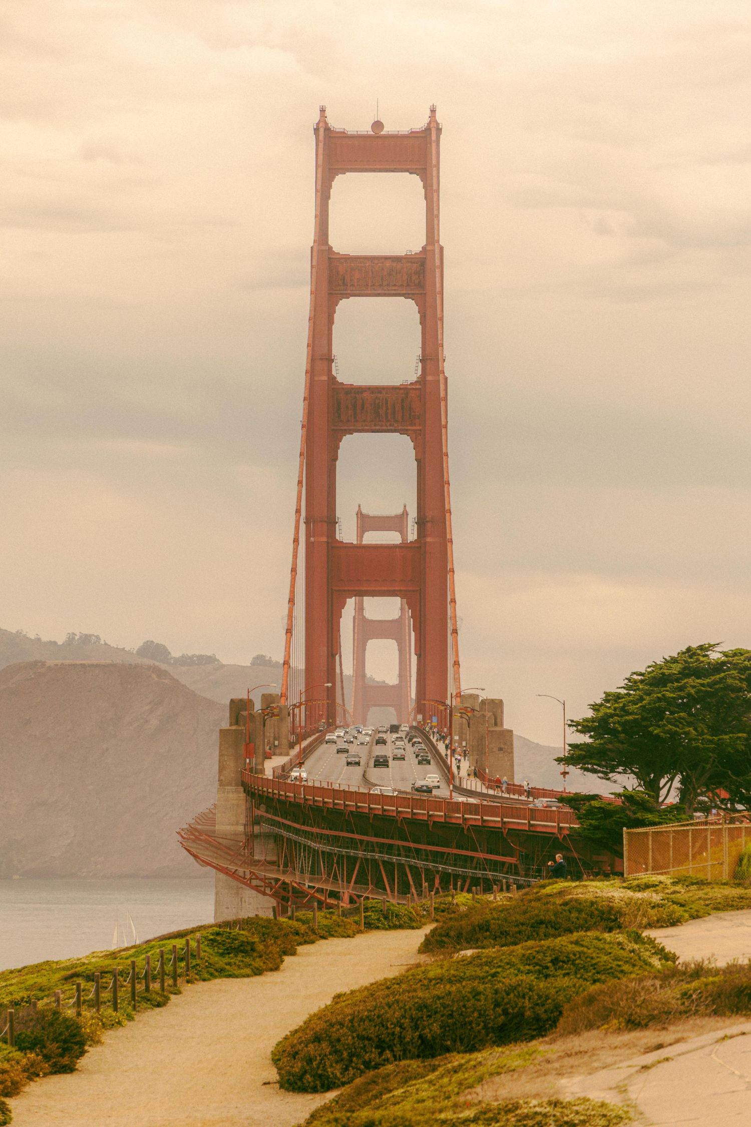 Golden Gate Bridge tower rises through misty fog with traffic below, viewed from a sandy path in San Francisco.