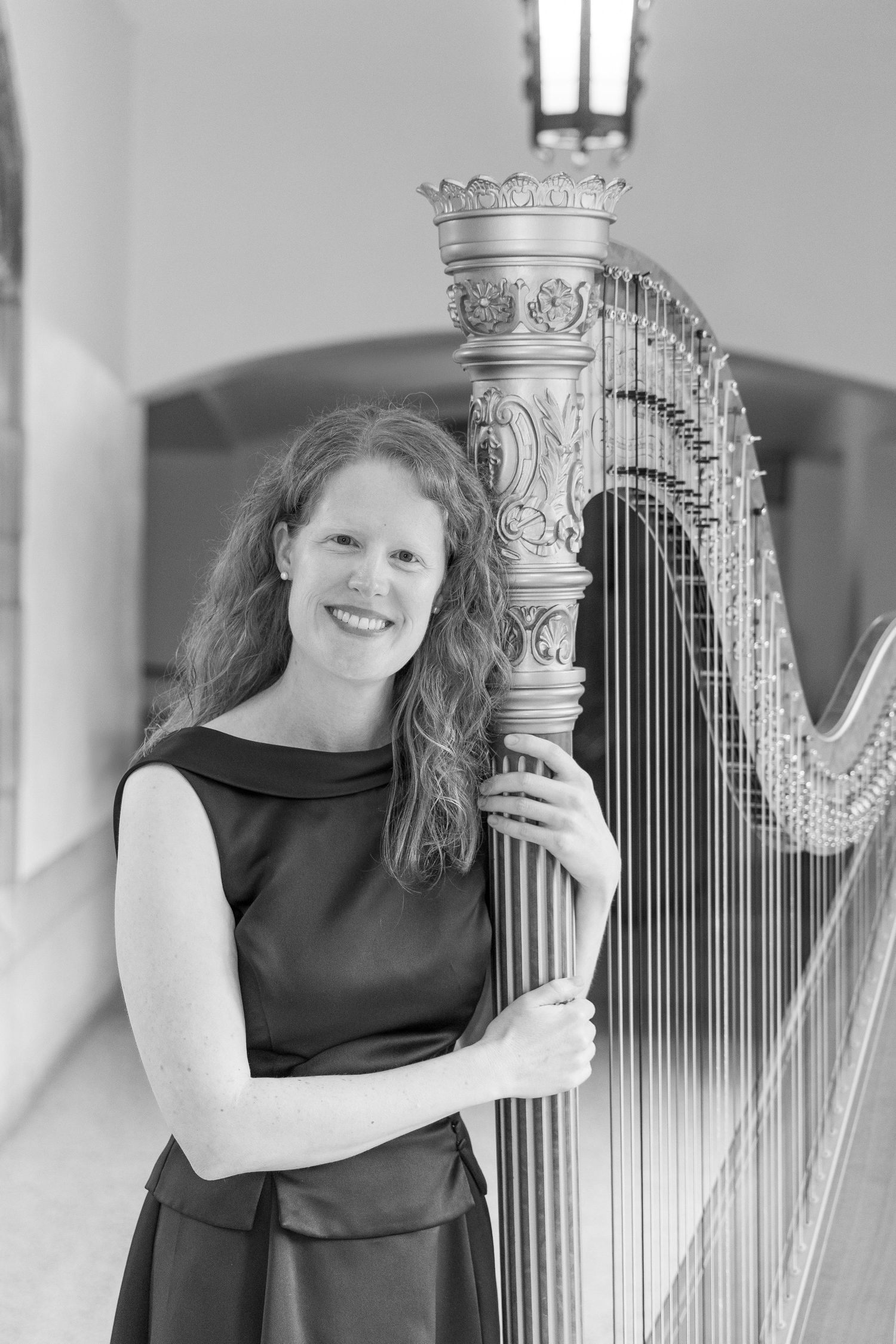 Woman smiling while posing with an ornate concert harp in an elegant arched hallway, black and white photo.