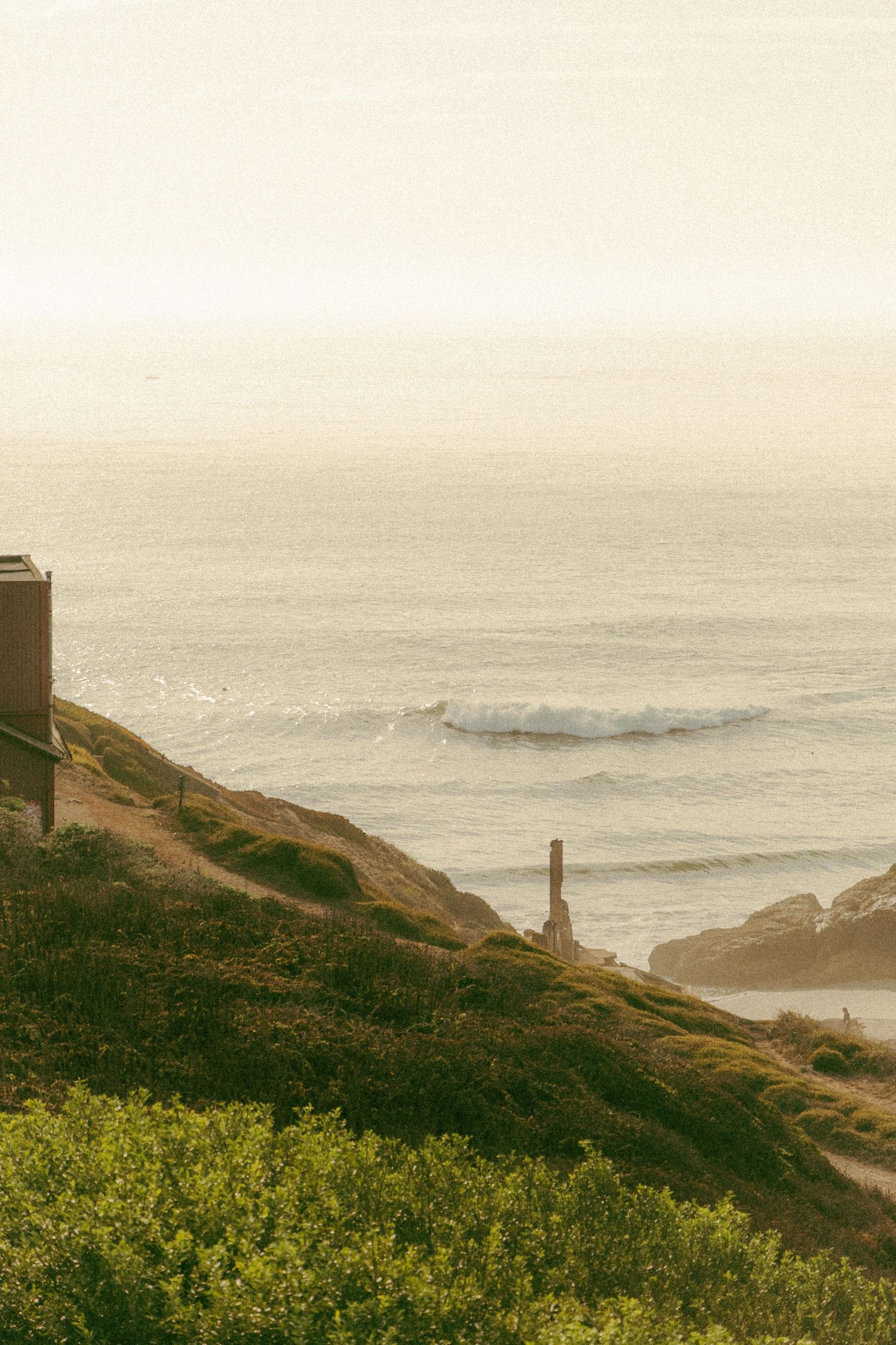 Misty coastal hillside with green vegetation overlooking ocean waves with building and wooden post at dusk.