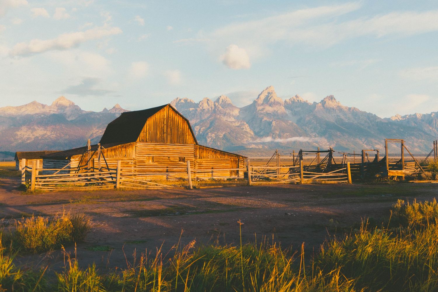 Rustic wooden barn at golden hour with Grand Teton mountain range backdrop in Wyoming's scenic landscape.