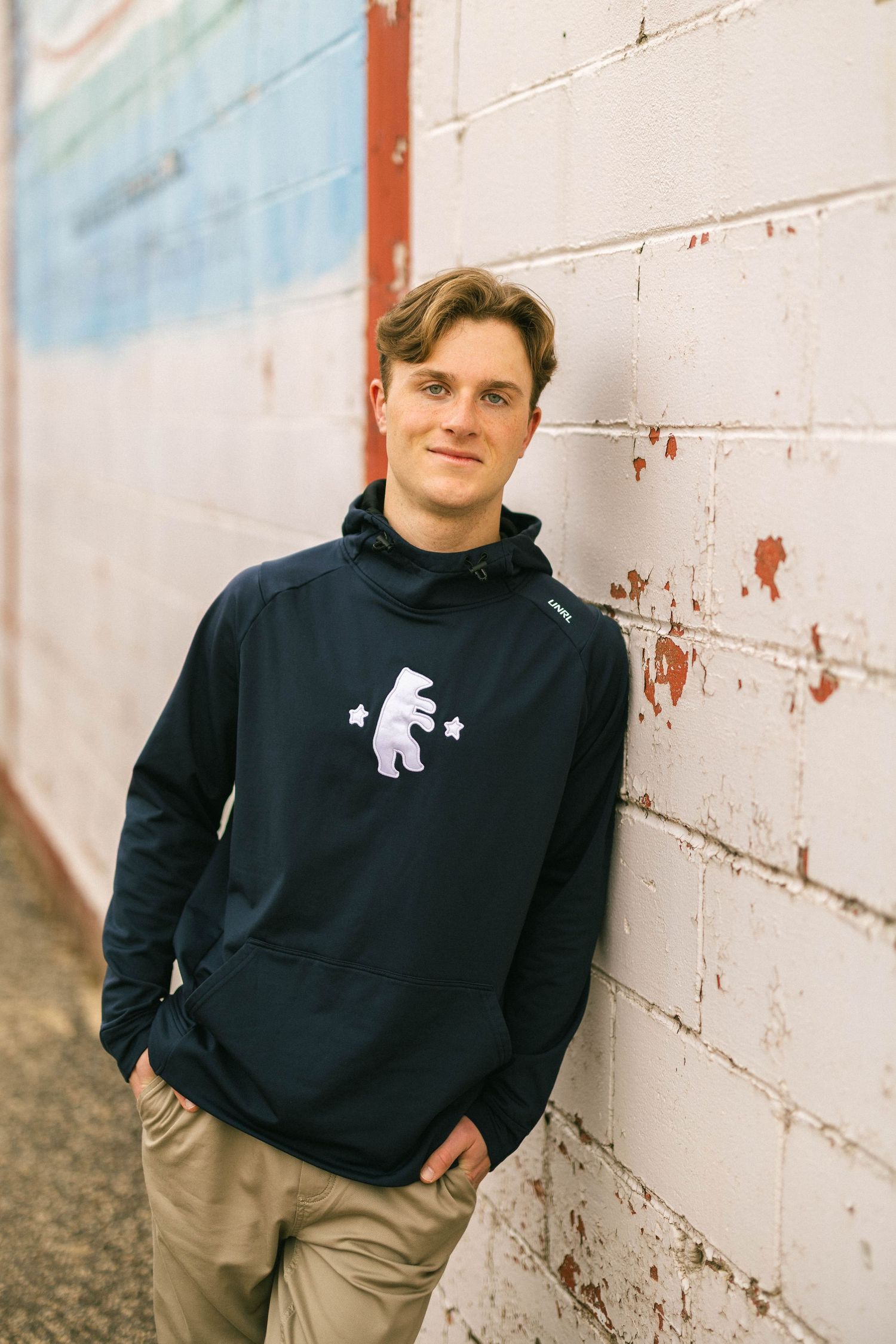 Young man in navy bear graphic hoodie leaning against white brick wall.