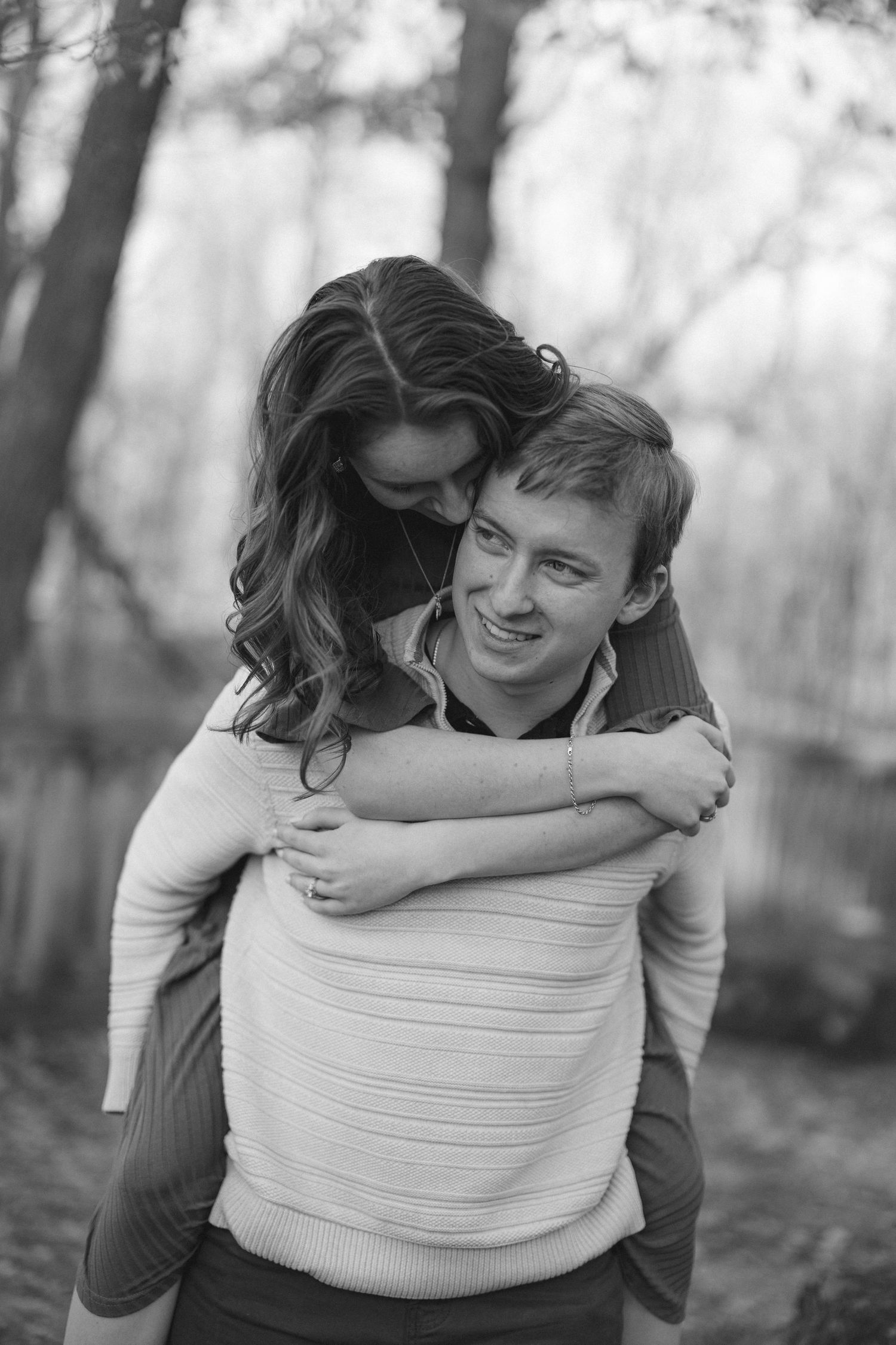 Black and white photo of smiling man giving woman a piggyback ride in a woodland setting.