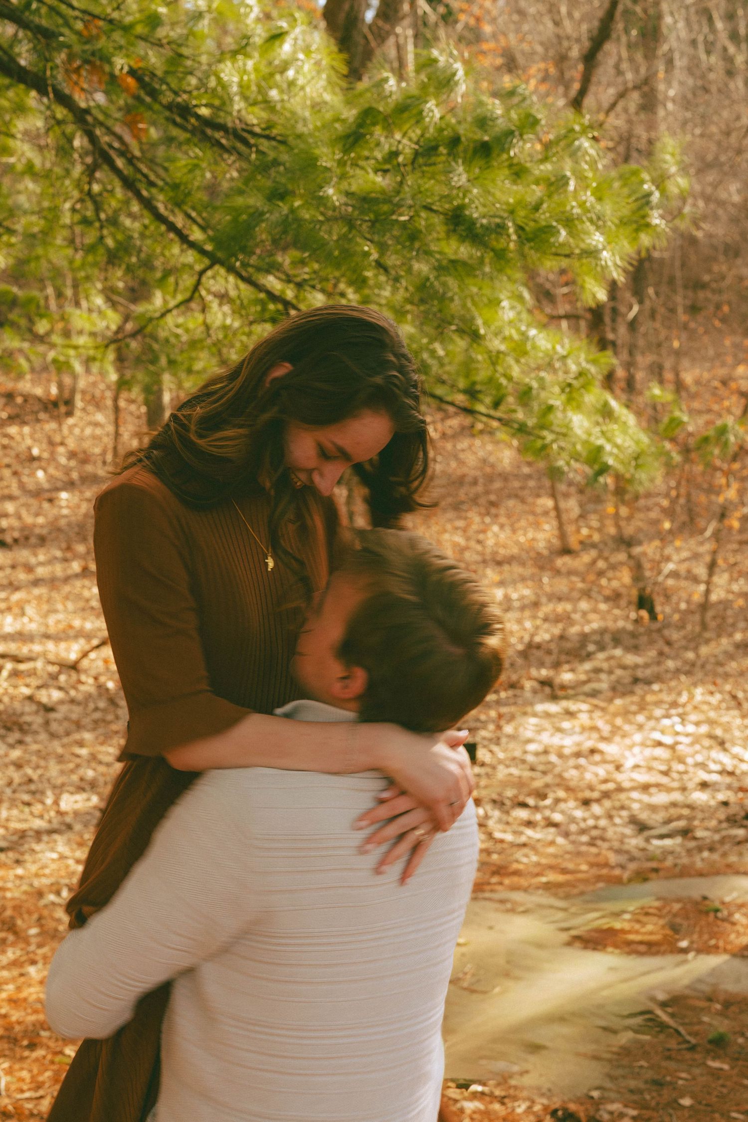 Woman laughing joyfully while hugging a man beneath green pine branches in a fall forest.