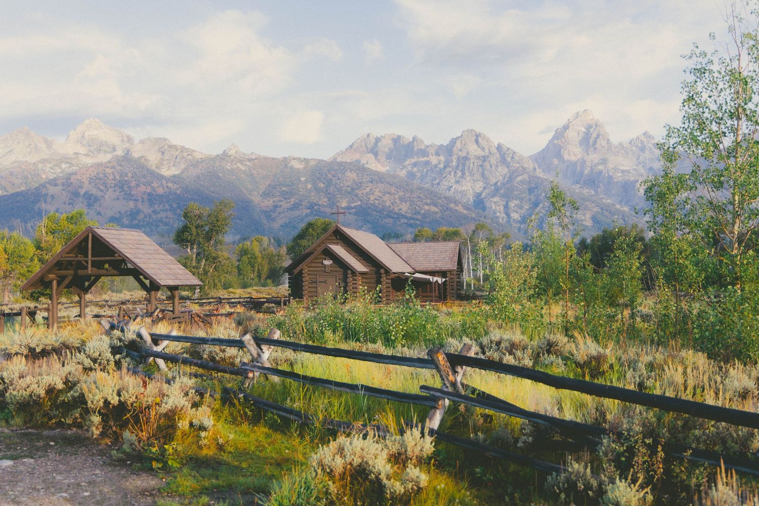 Rustic wooden cabins nestled in a meadow with the Grand Teton mountain range in the background under a cloudy sky.