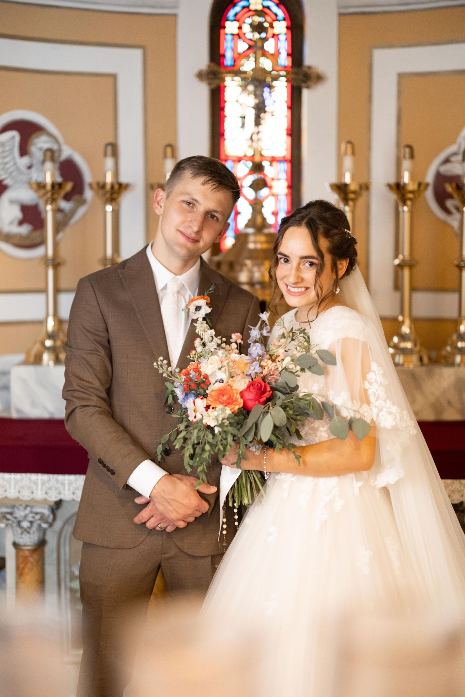 Bride and groom pose together at church altar holding a colorful bridal bouquet of roses and wildflowers.