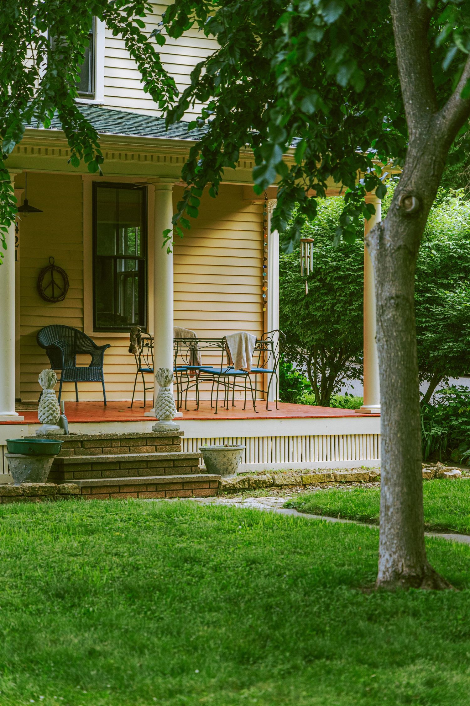 Yellow farmhouse with covered porch featuring black metal chairs surrounded by lush green summer trees and manicured lawn.
