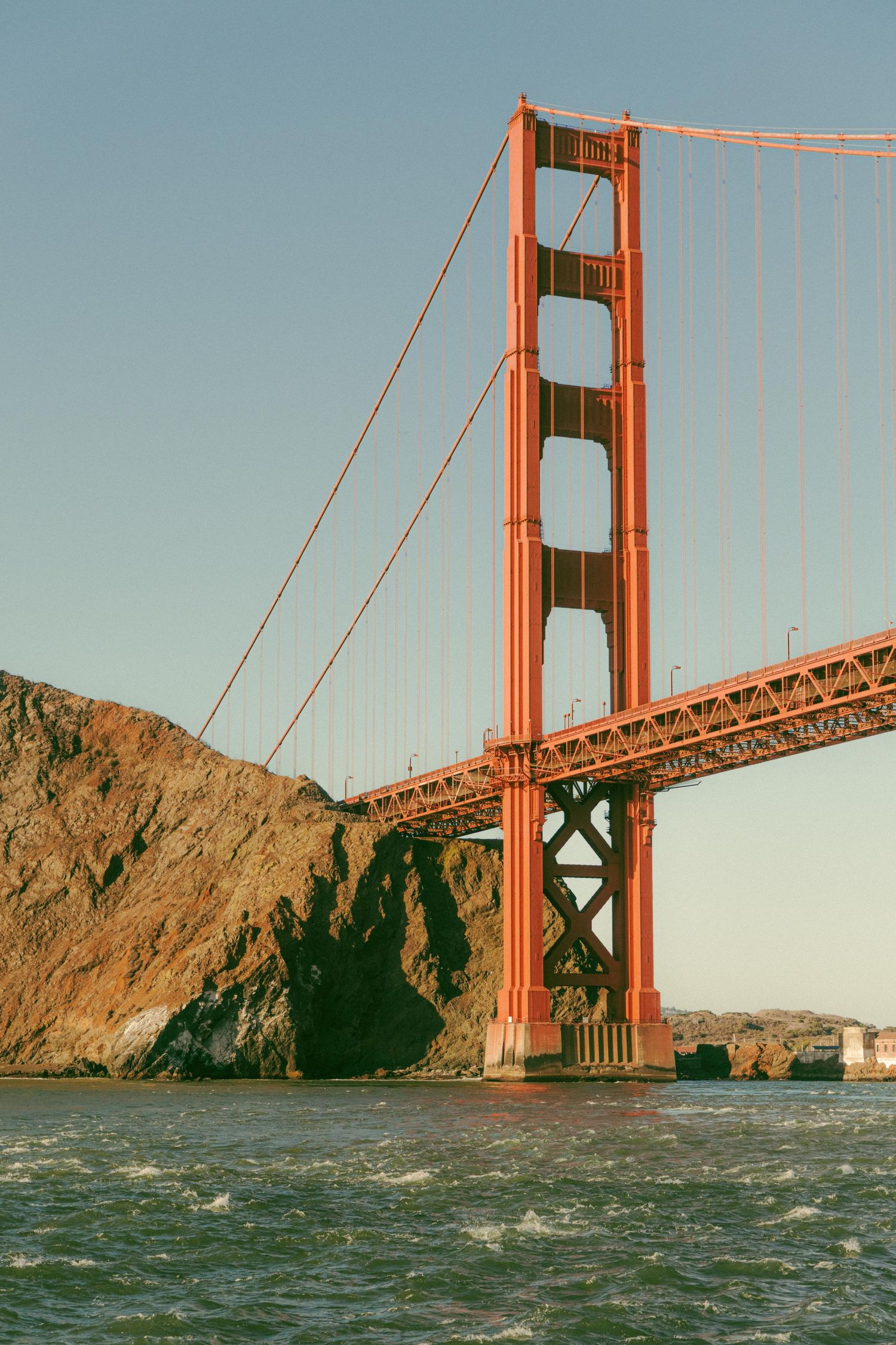 Golden Gate Bridge red tower rising above rocky San Francisco Bay coastline at golden hour sunset.
