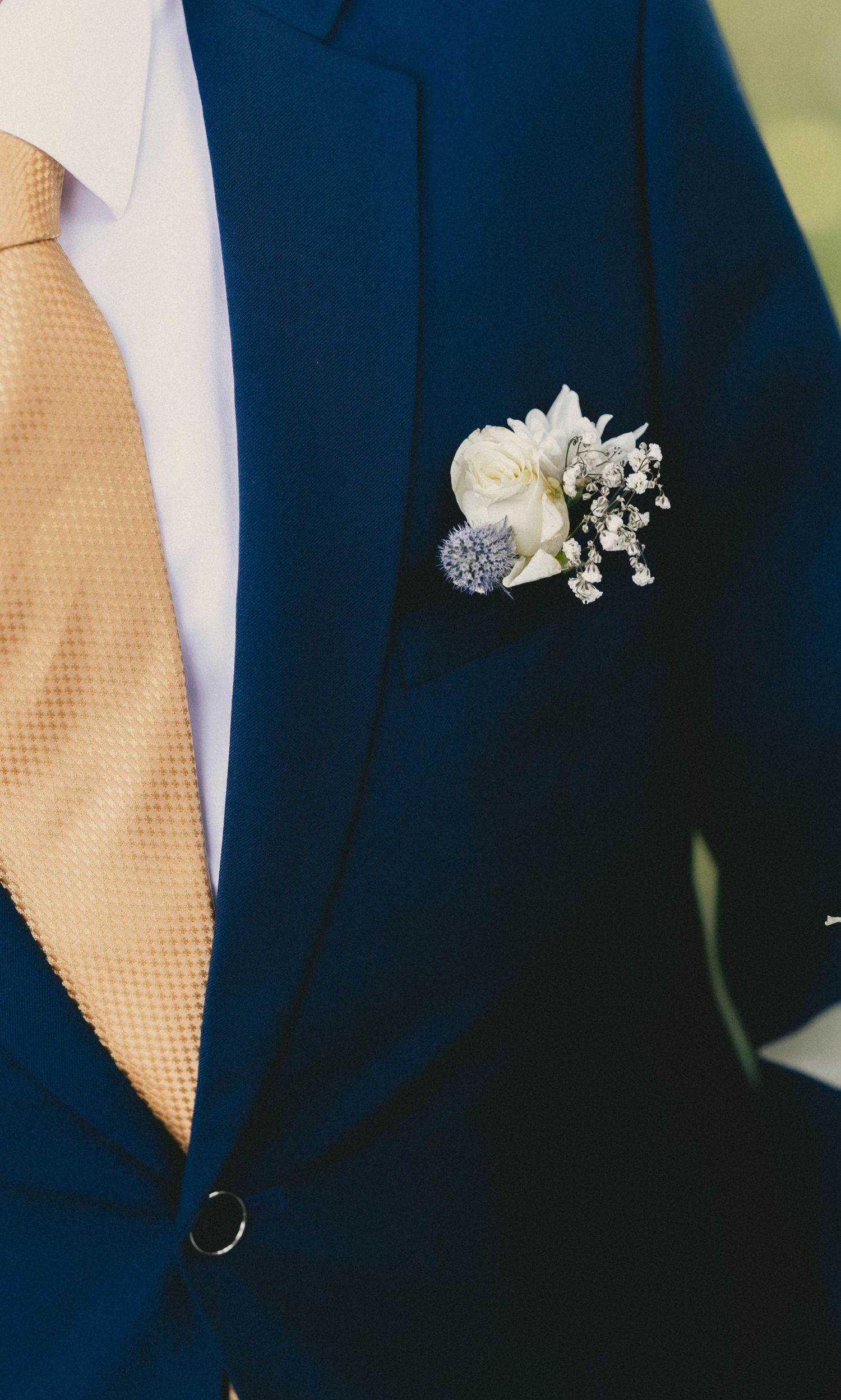A groom in a navy blue suit with a gold tie and white floral boutonniere featuring baby's breath and a blue thistle.