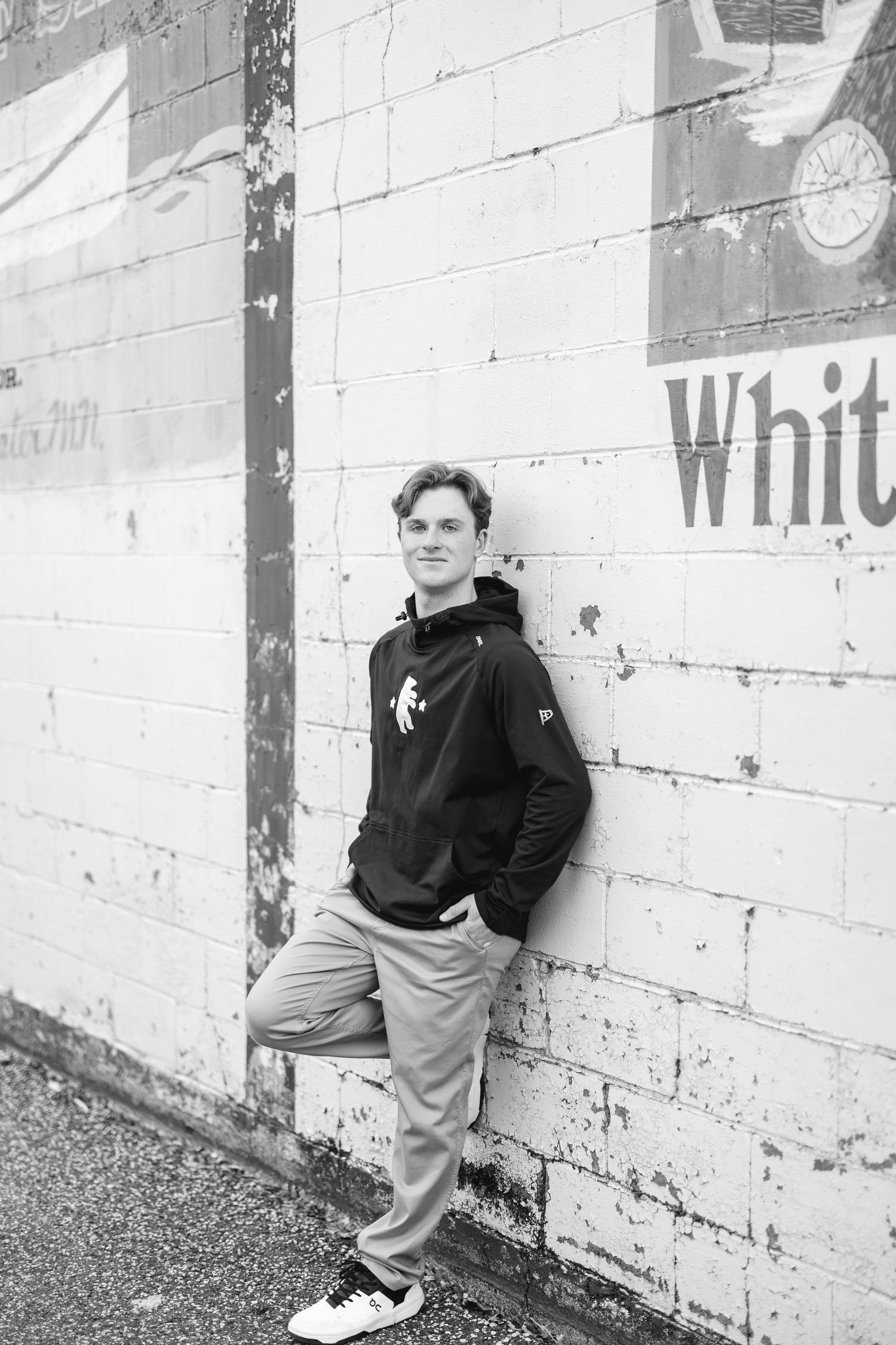 Black and white photo of a young man in a hoodie leaning against a brick wall with faded signage.