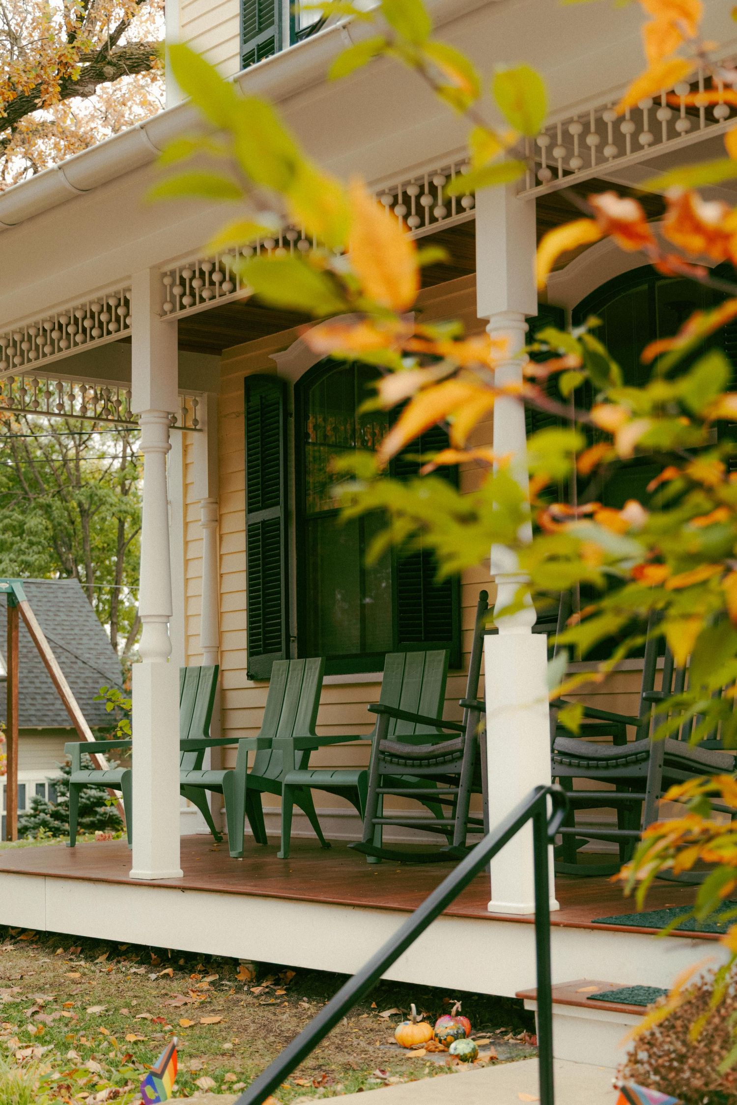 Charming white Victorian home porch with green rocking chairs, decorative trim, and colorful autumn foliage in foreground.