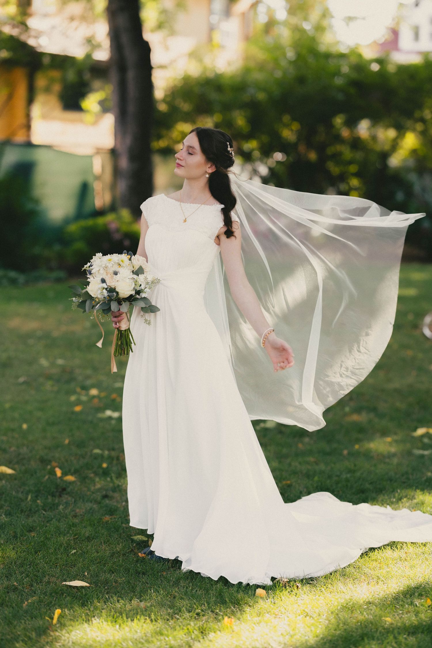 Bride in white gown with flowing veil holding bouquet outdoors on green lawn.