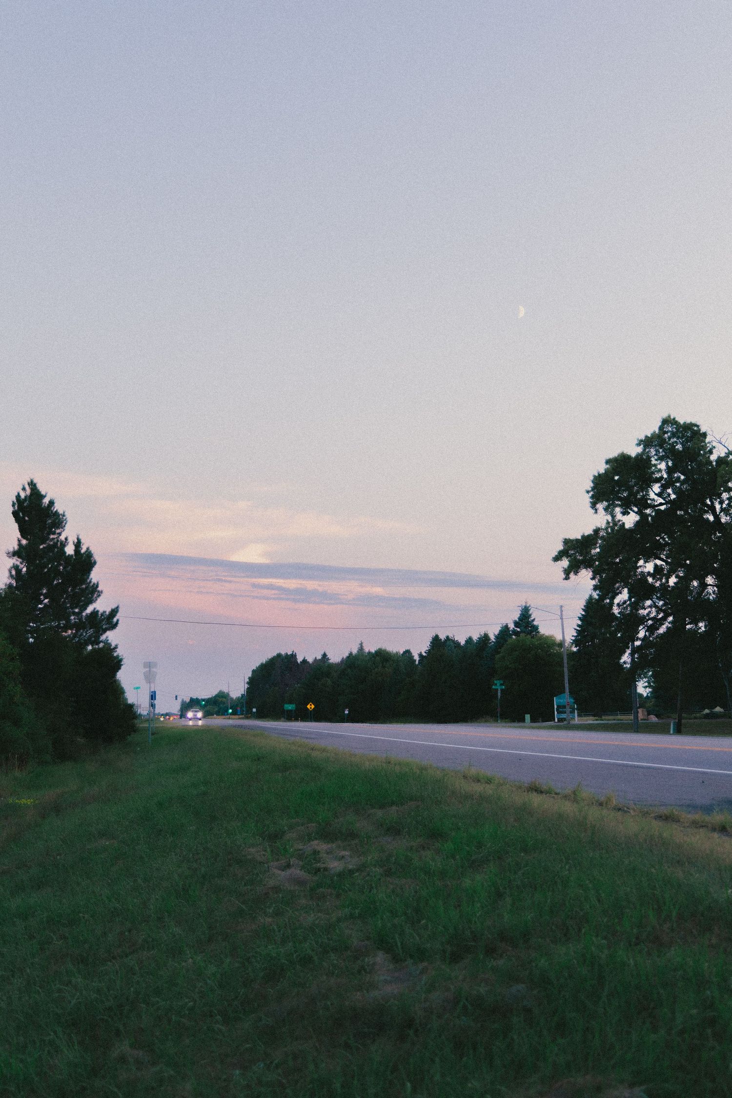 Twilight dusk sky over a quiet rural highway lined with lush green trees and grass, with a crescent moon visible.