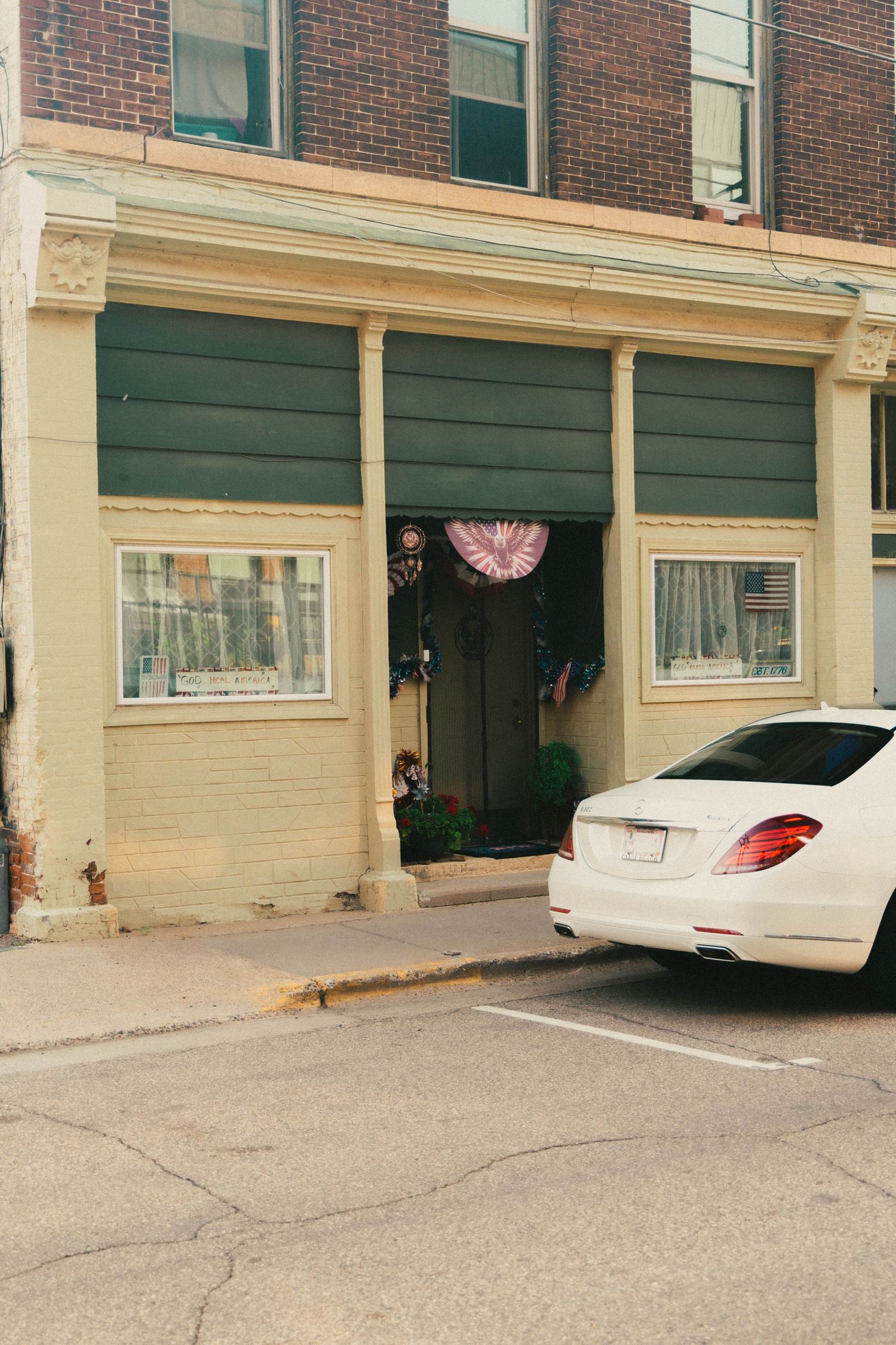 White Mercedes sedan parked outside a brick building with green awning and patriotic decorations on a quiet street.