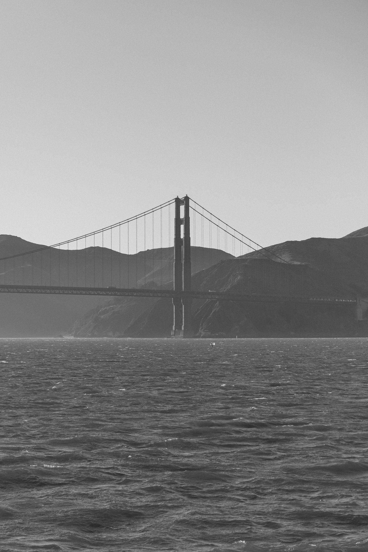 Black and white panoramic view of the Golden Gate Bridge spanning the bay with misty hills in the background.