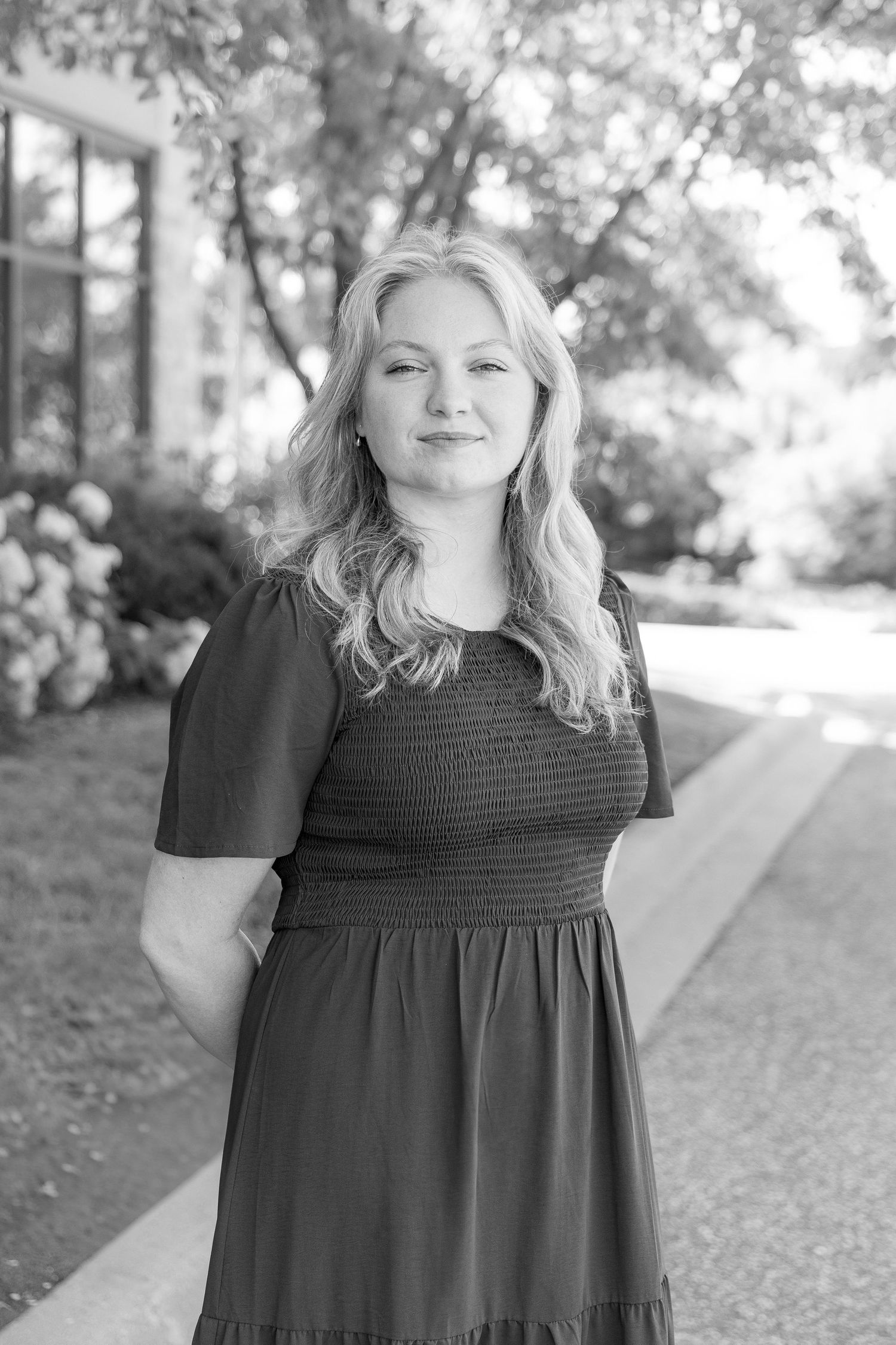 Black and white portrait of a young blonde woman in a smocked dress posing outdoors near flowering bushes.