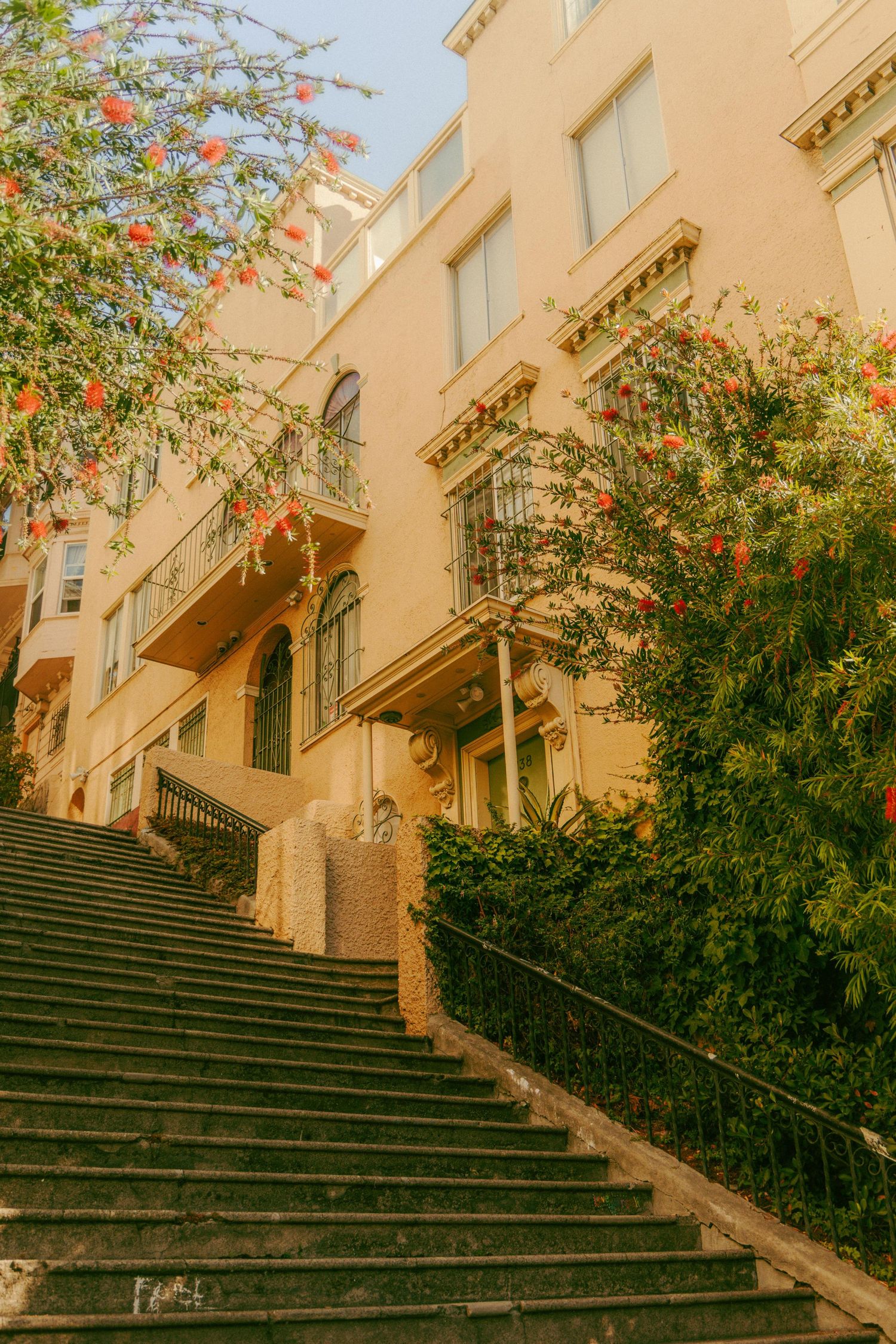 Stone staircase leading up to a yellow Mediterranean style building adorned with blooming red flowers and lush greenery.