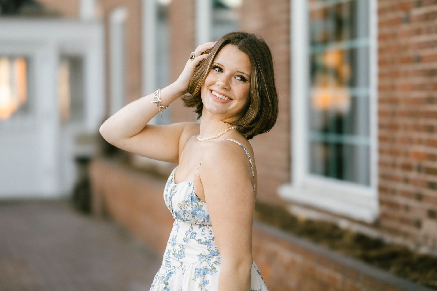 Young woman in floral sundress smiling over shoulder, posing outdoors near brick building.