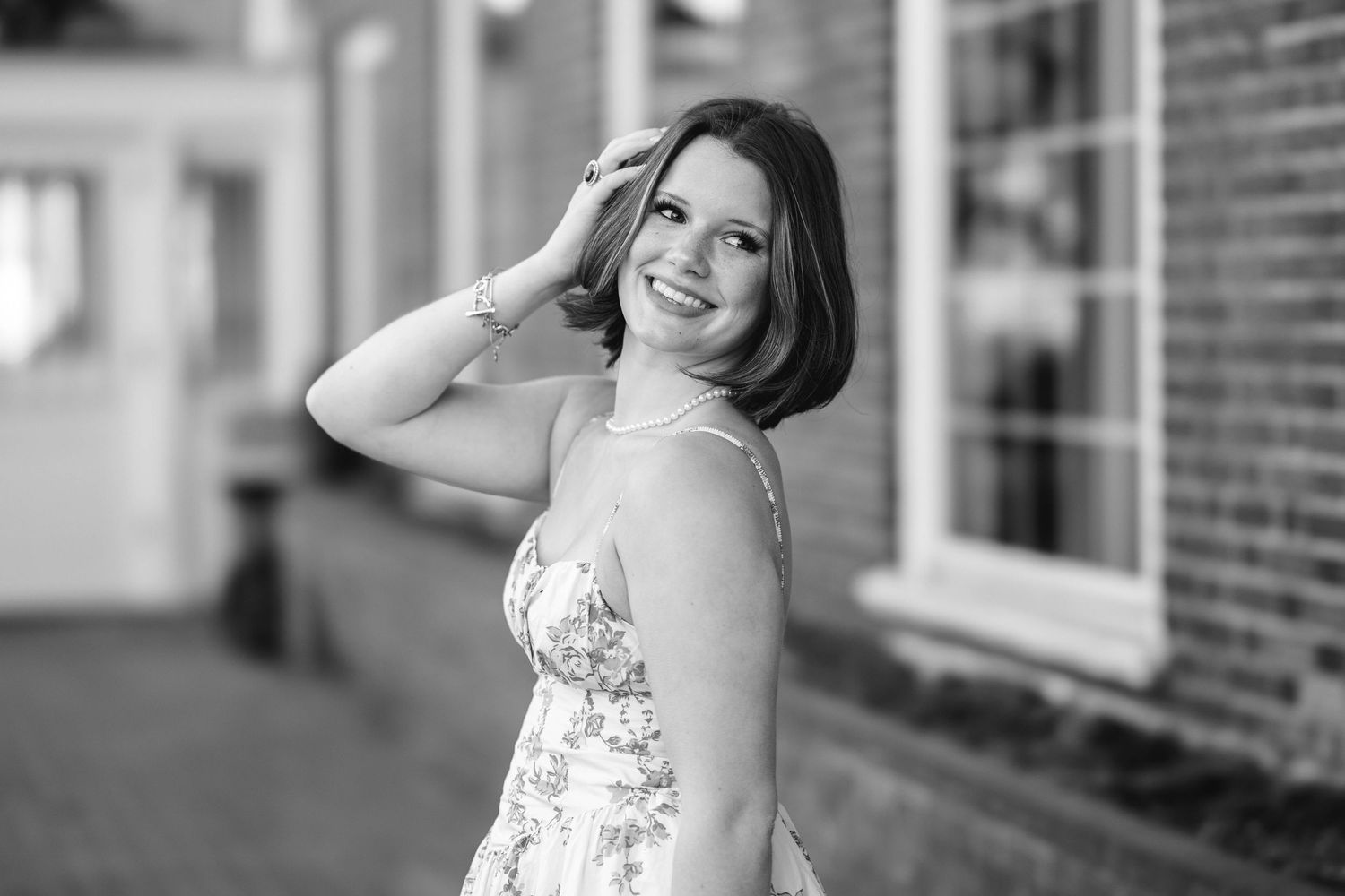 Black and white portrait of a smiling young woman in a floral dress posing outdoors near a brick building.
