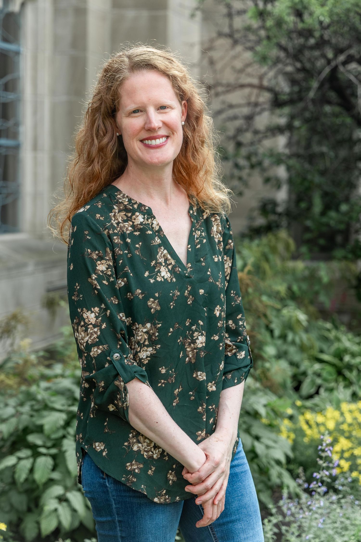 Happy redhead woman in green floral blouse smiling in lush garden beside stone building.