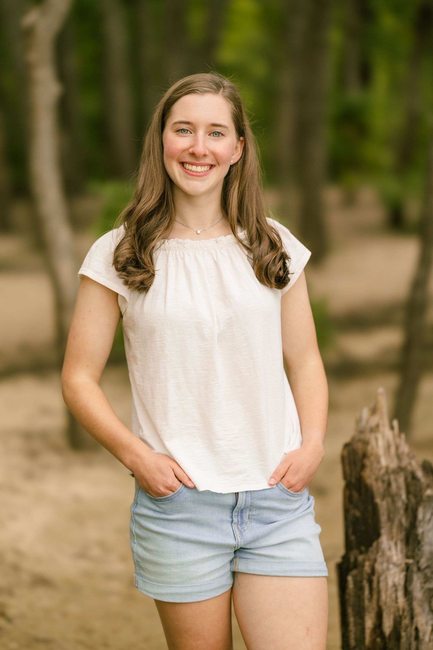 Smiling teenage girl in white top and denim shorts posing outdoors in a wooded setting for senior portraits.