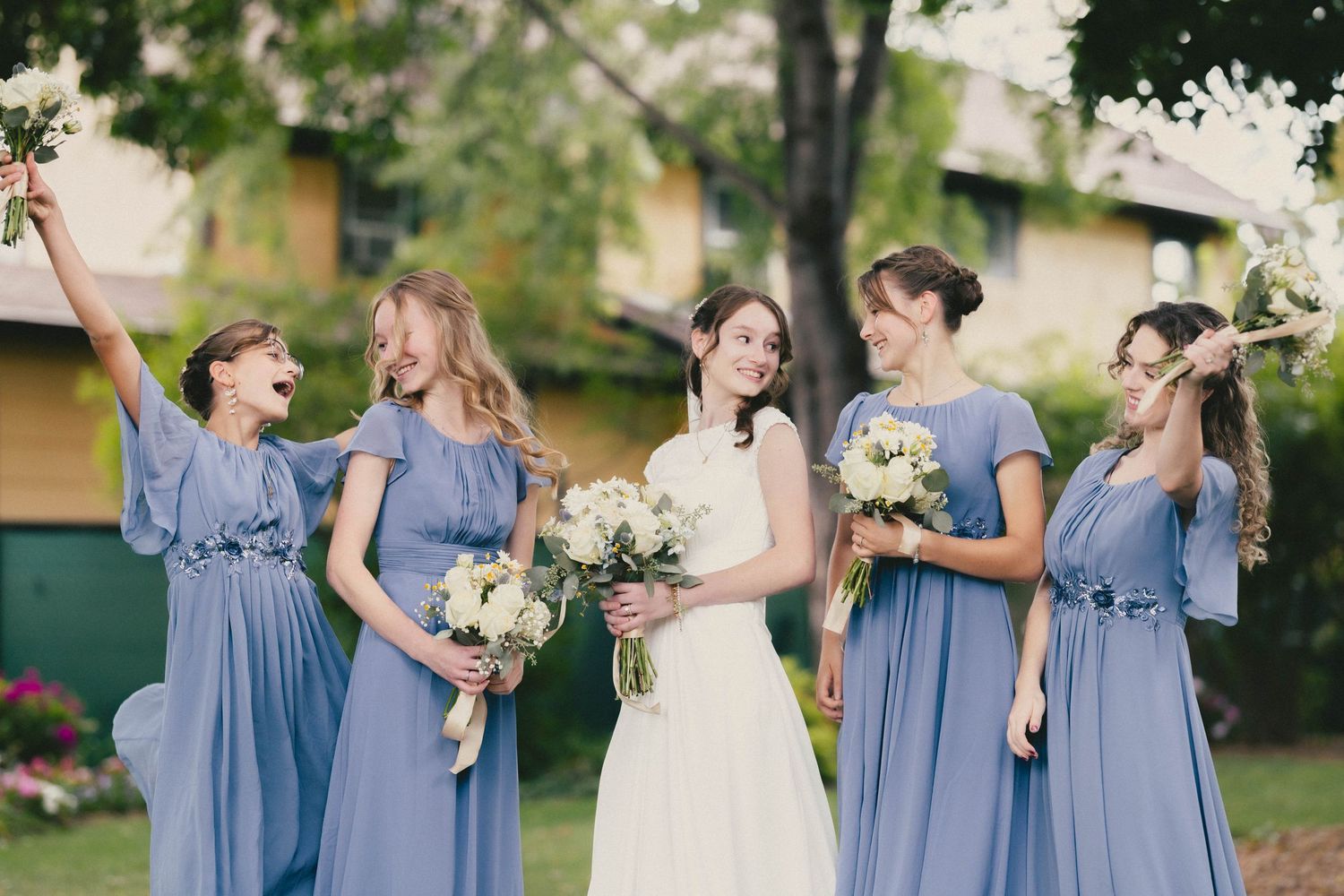 Bride in white gown poses with five bridesmaids in blue-gray dresses holding bouquets outdoors.