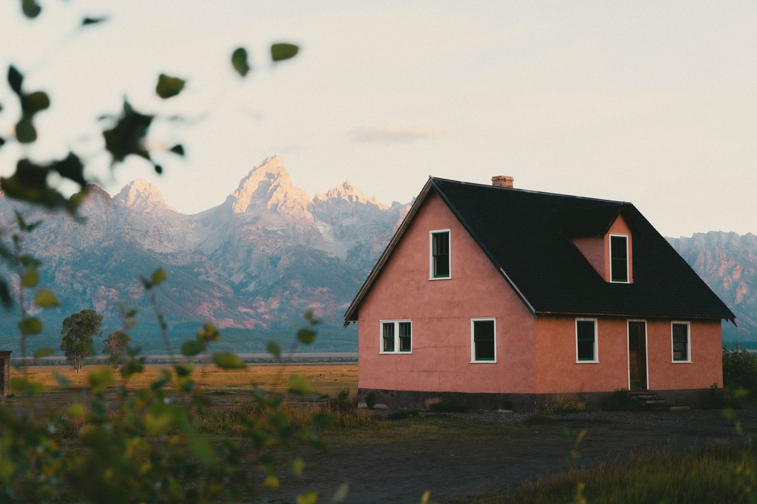 Pink farmhouse with black roof sits against dramatic snow-capped Grand Teton mountains at dusk.