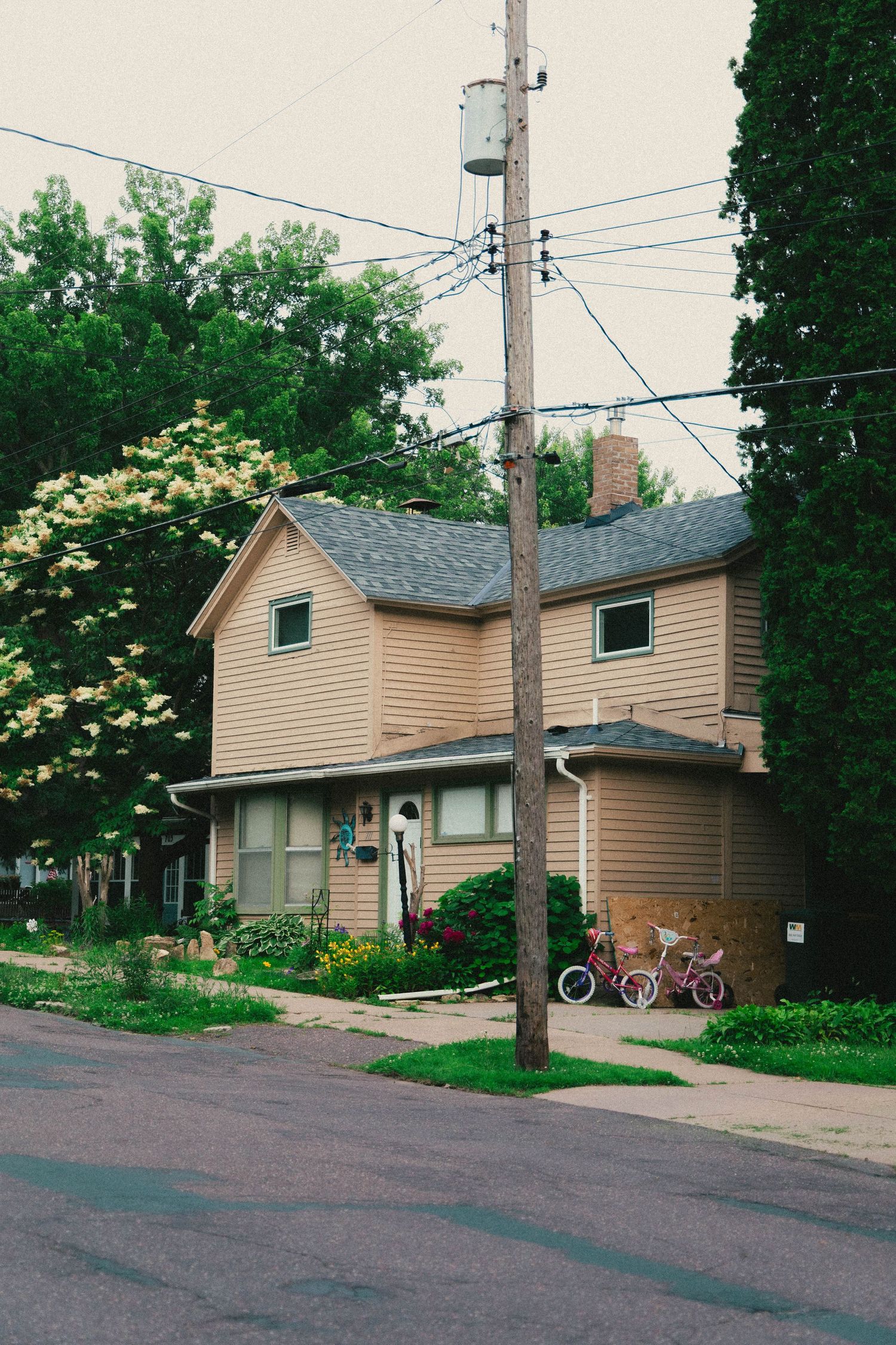 A yellow two-story house with a utility pole, power lines, bicycles, and lush green trees on a quiet residential street.