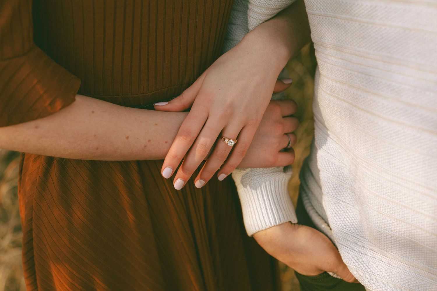 Close-up of woman's hand with gold engagement ring resting on partners arm outdoors.