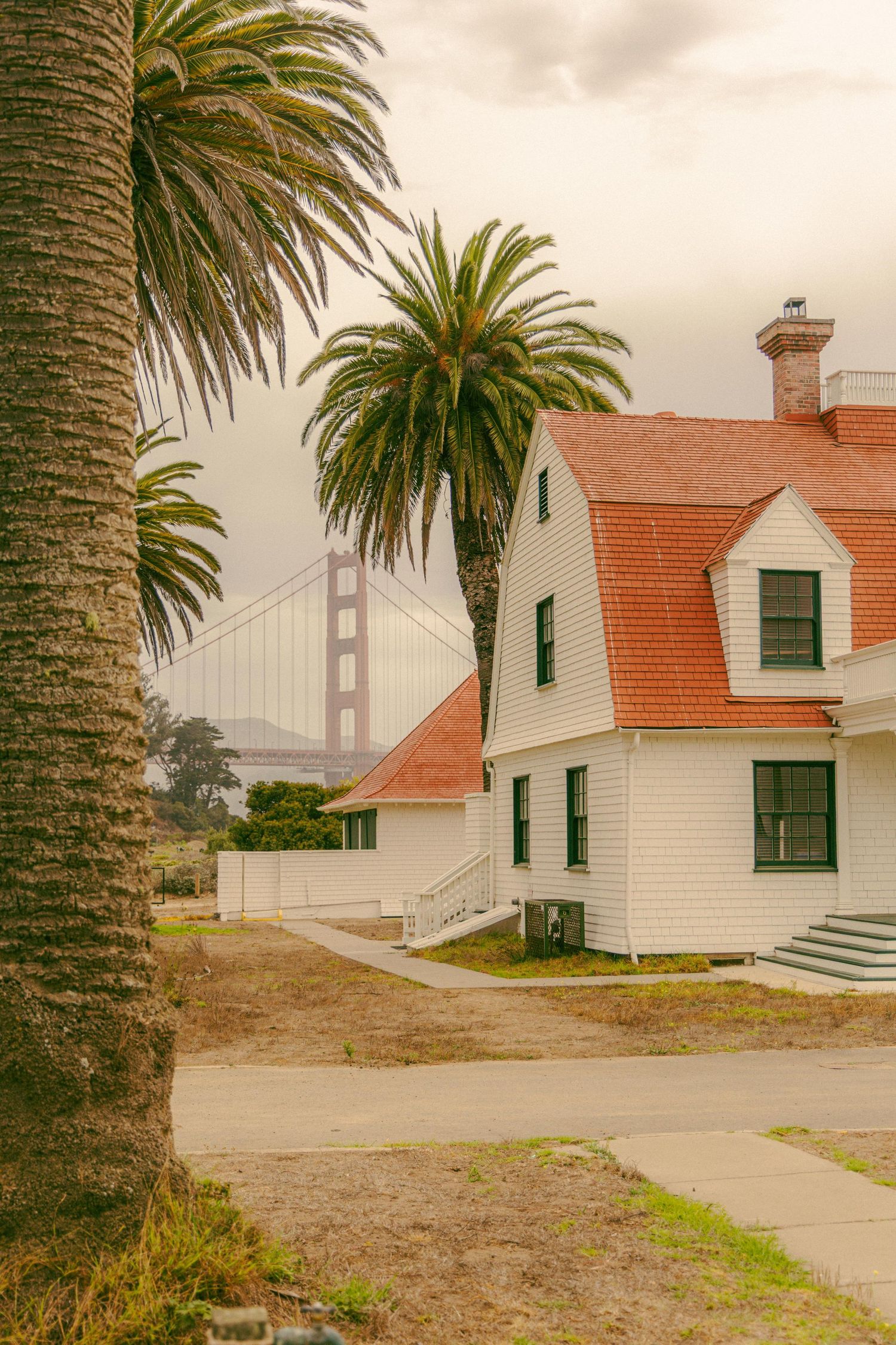 Historic white cottage with red roof near the Golden Gate Bridge partially obscured by fog beside a tall palm tree.